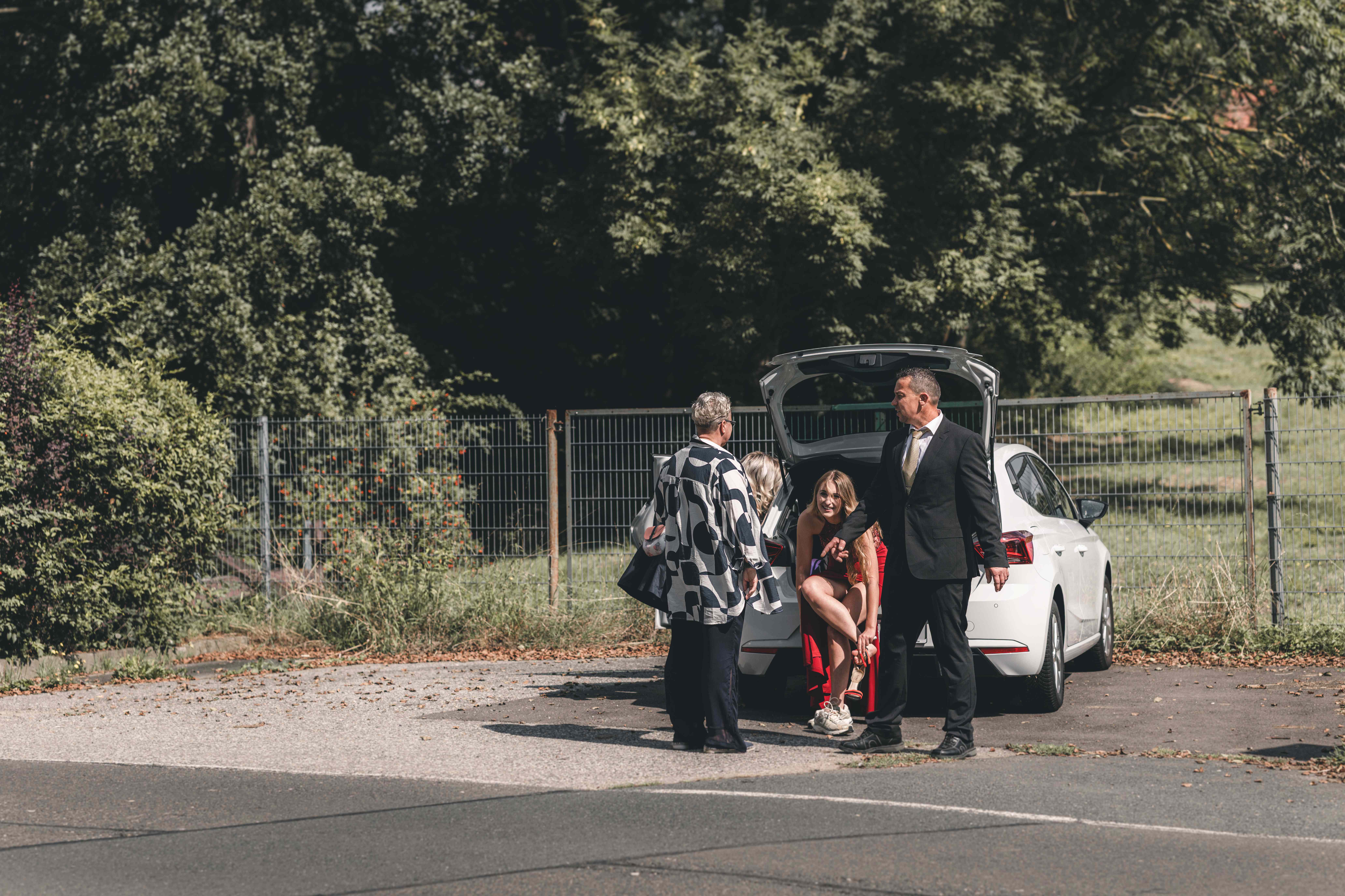 Hochzeitsgäste und Helfer stehen im Freien an der geöffneten Heckklappe eines weißen Autos auf dem Parkplatz vor der Kirche.