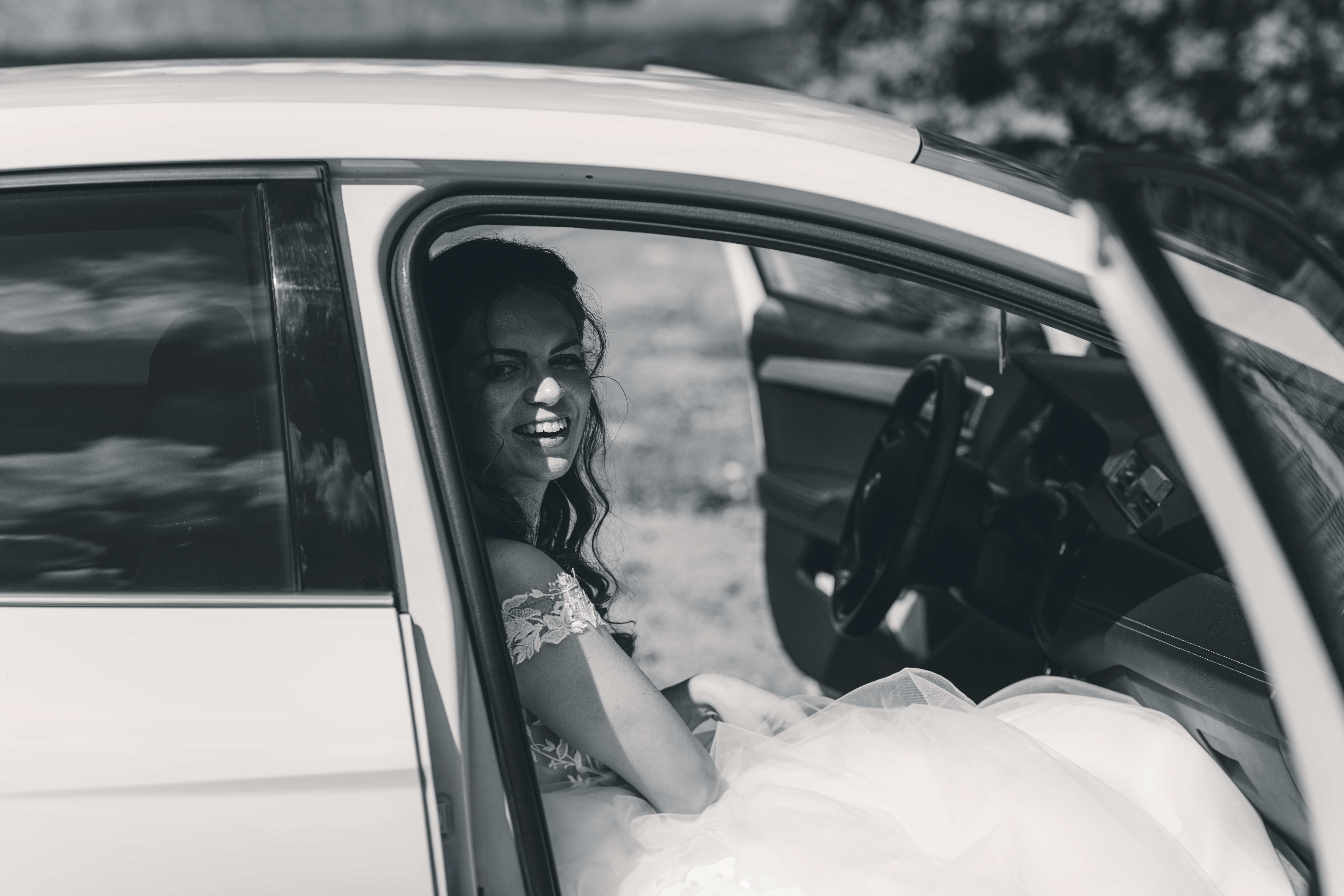 Eine stimmungsvolle Schwarz-Weiß-Aufnahme der Braut im Spitzenkleid, die kurz vor der kirchlichen Trauung glücklich lächelnd aus dem Auto blickt.