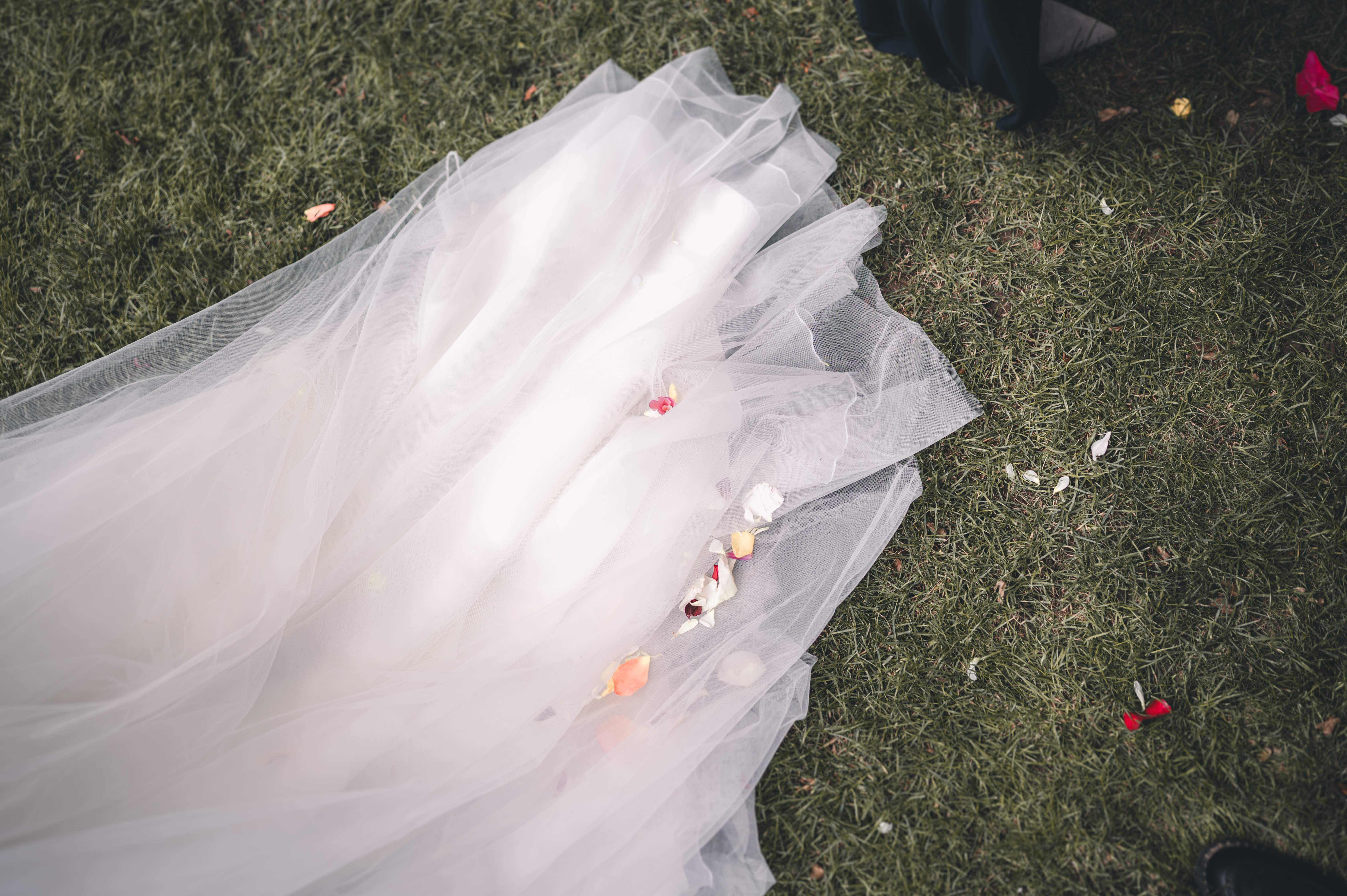 Nahaufnahme der weißen Tüllschleppe des Brautkleides auf grünem Gras, auf der bunte Blütenblätter nach den Gratulationen liegen.