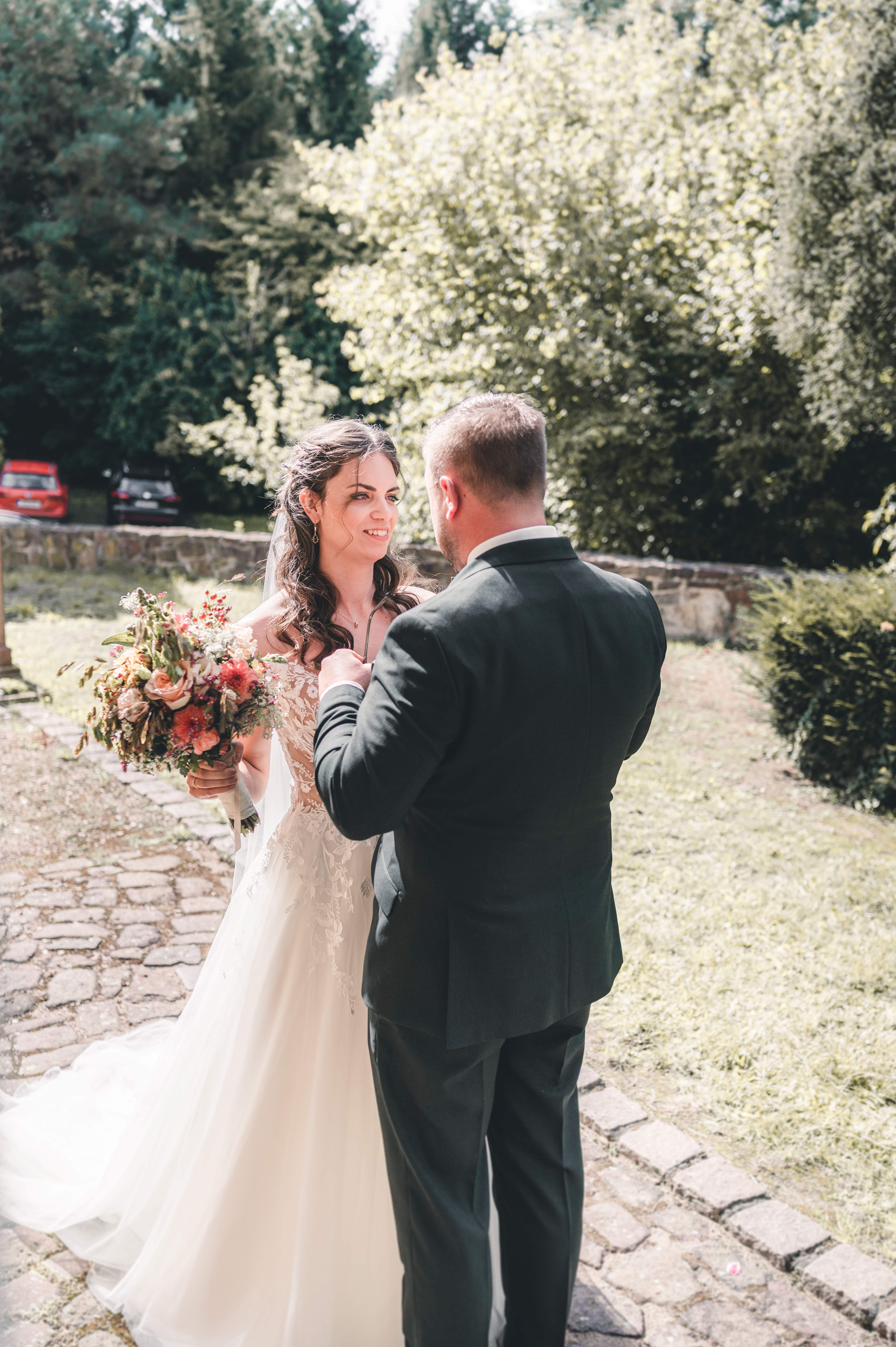 Die Braut im weißen Hochzeitskleid steht lächelnd mit ihrem Wiesenblumenstrauß neben ihrem Bräutigam auf dem gepflasterten Kirchweg.