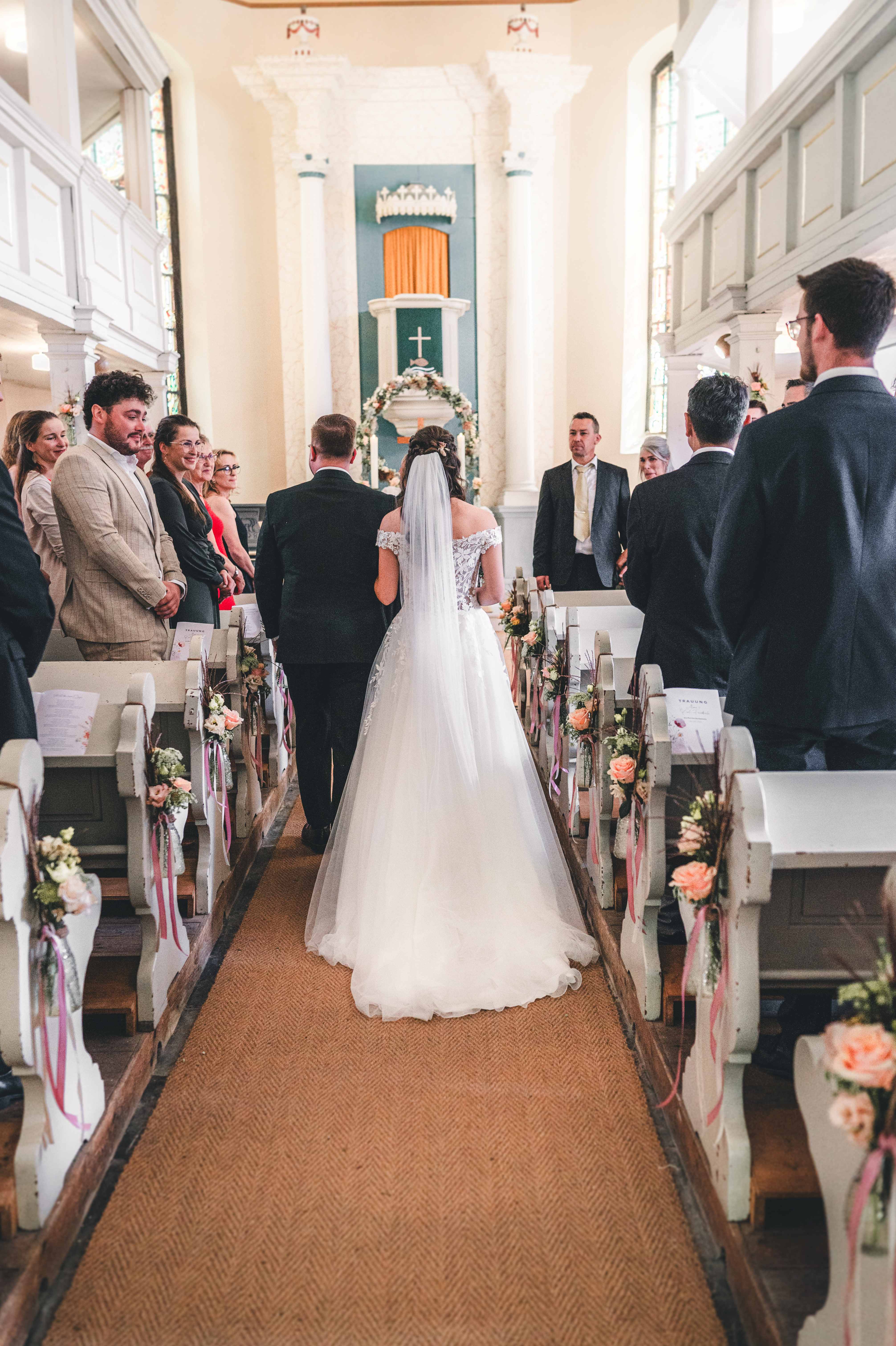 Rückansicht des Brautpaares beim Einzug durch den Mittelgang der Kirche, vorbei an Gästen und mit frischen Blumen geschmückten Kirchenbänken.