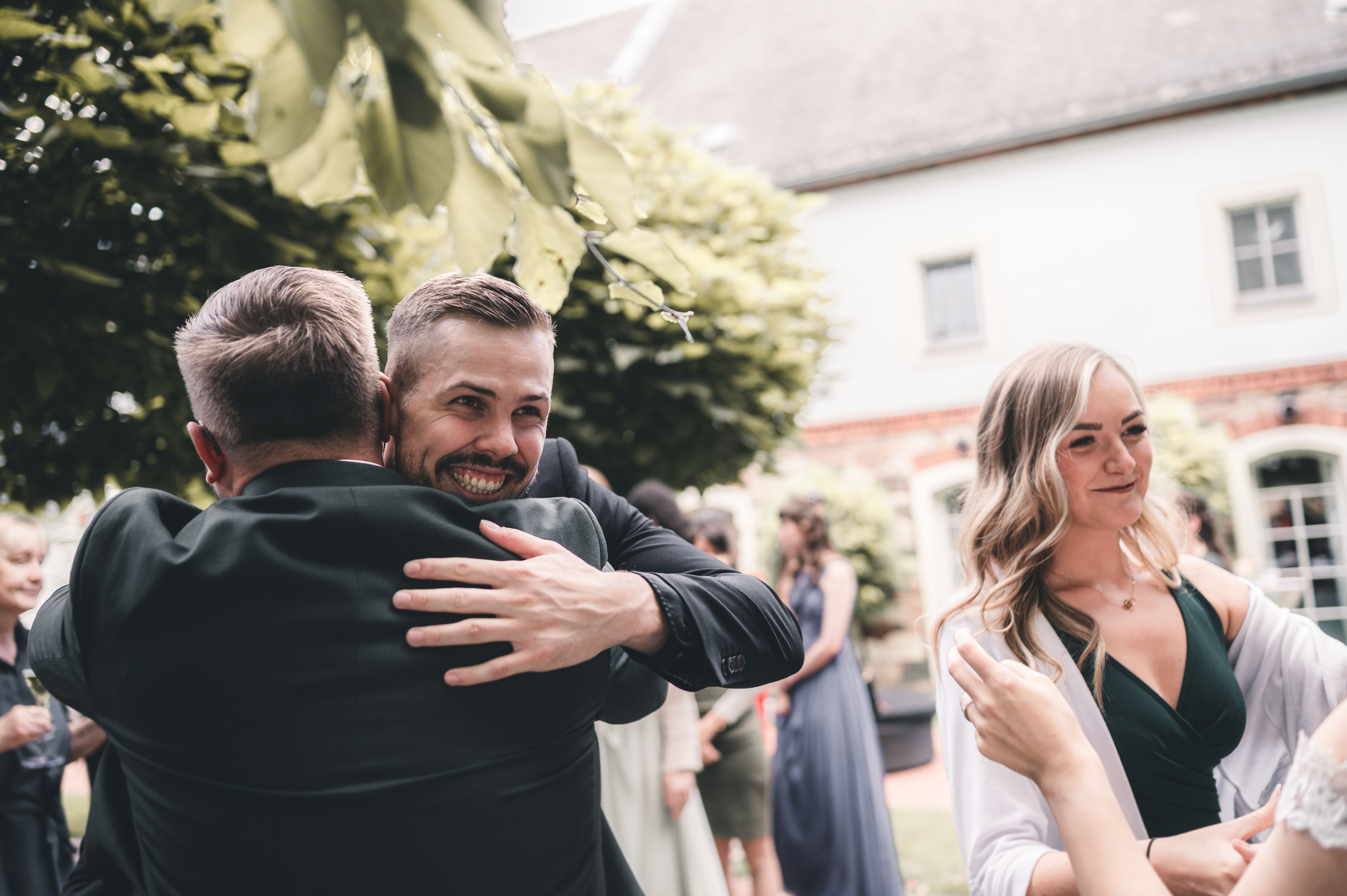 Der Bräutigam lacht herzlich, während er von einem guten Freund im Außenbereich der Location in Leisnig zur Hochzeit umarmt wird.