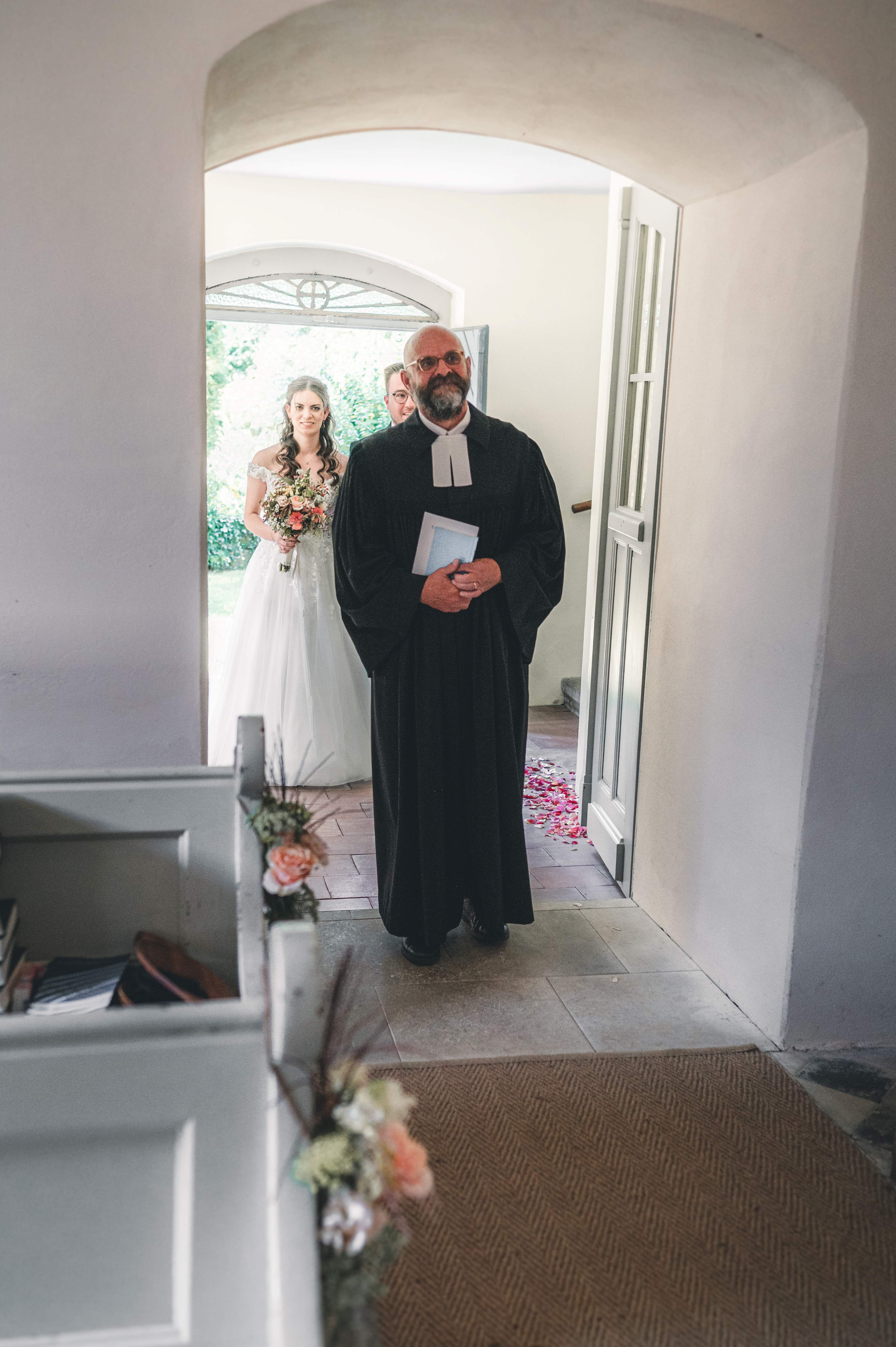 Nahaufnahme des Pfarrers, der das Brautpaar beim feierlichen Einzug durch den steinernen Torbogen in den Trausaal der Kirche führt.