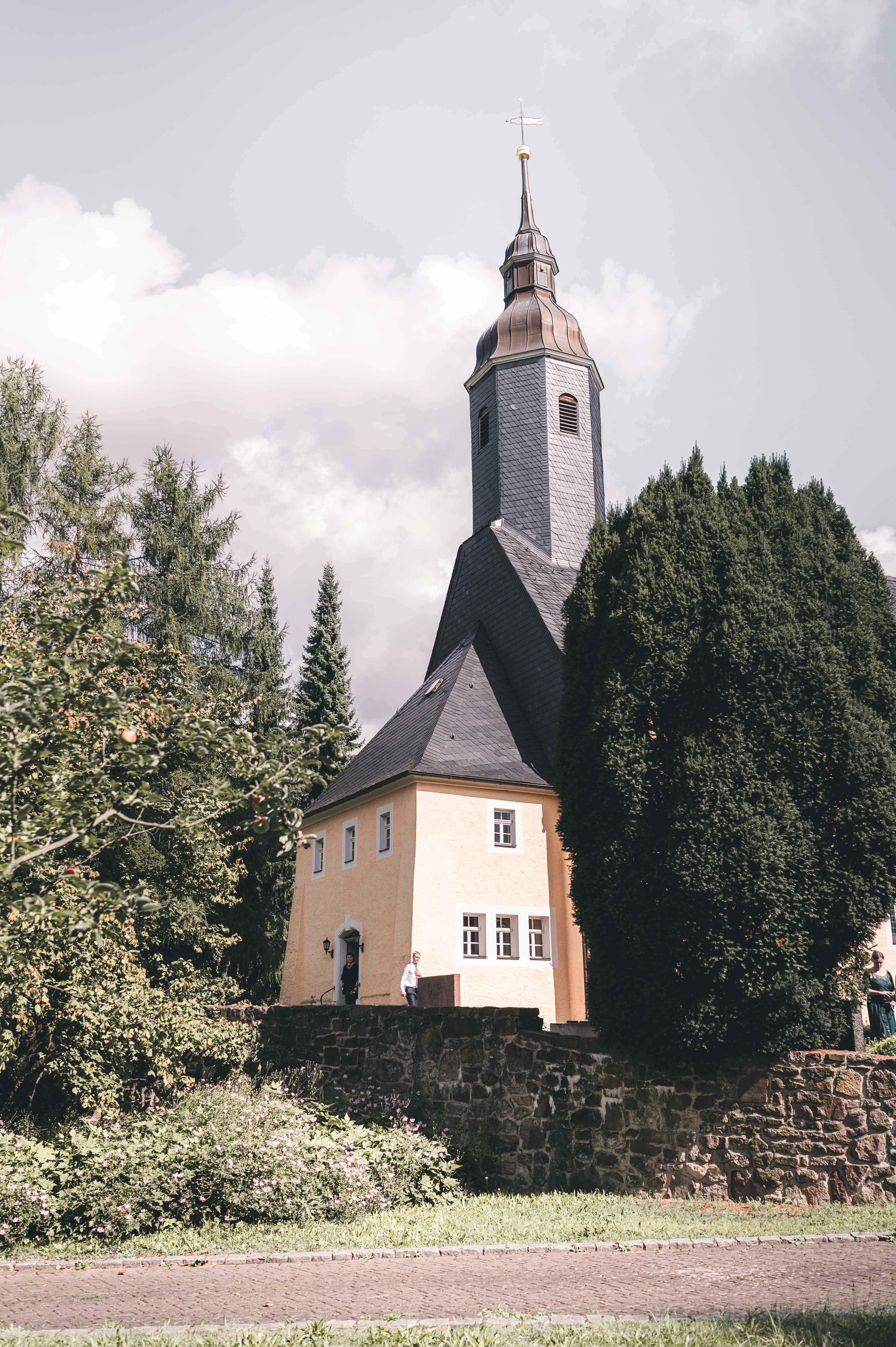 Außenansicht der gelb gestrichenen Kirche in Leisnig hinter einer alten Steinmauer, umgeben von dichten grünen Bäumen.