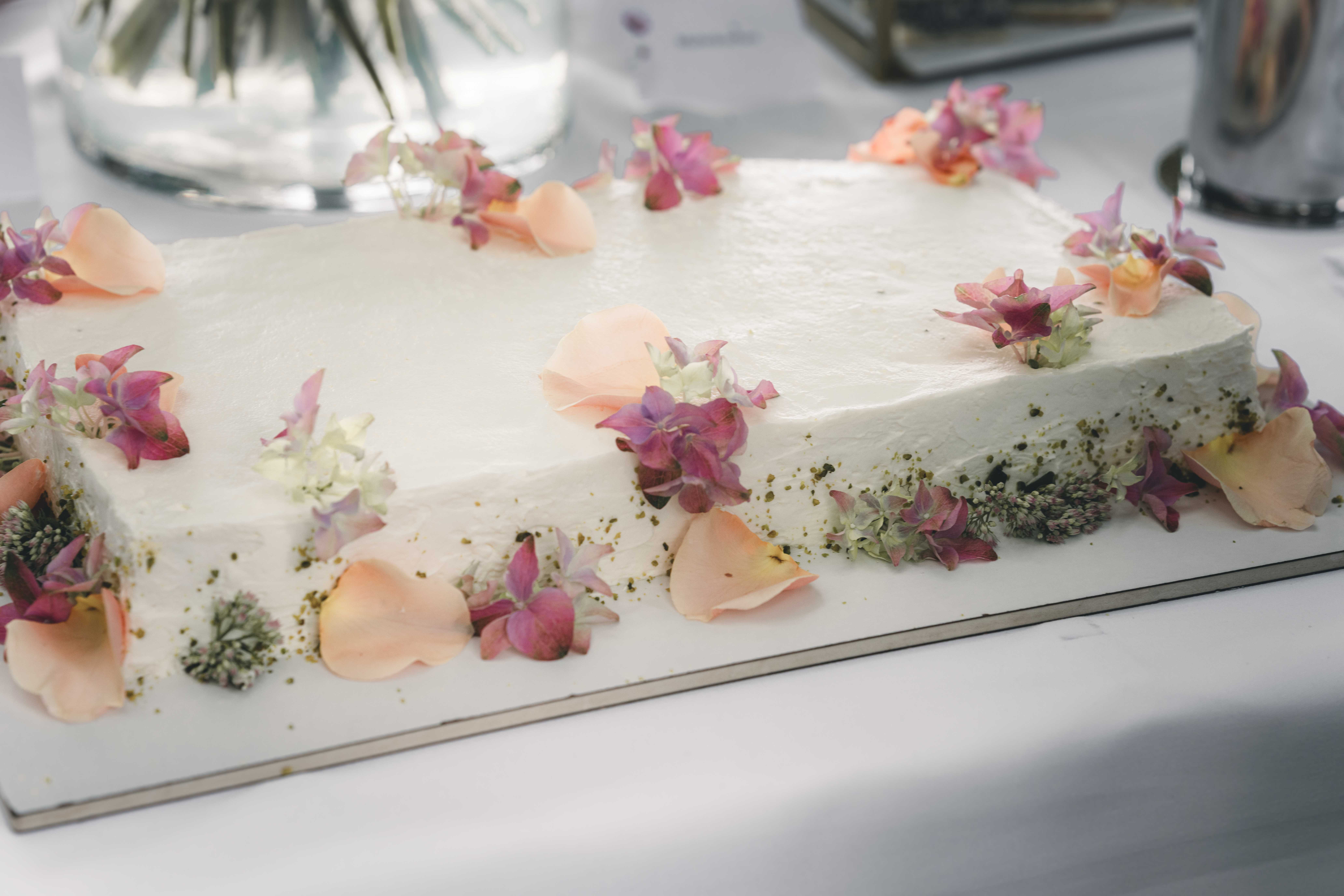 Eine elegant gestaltete, rechteckige Hochzeitstorte mit weißem Cremeflaschen-Design, verziert mit zarten Blütenblättern und gehackten Pistazien.