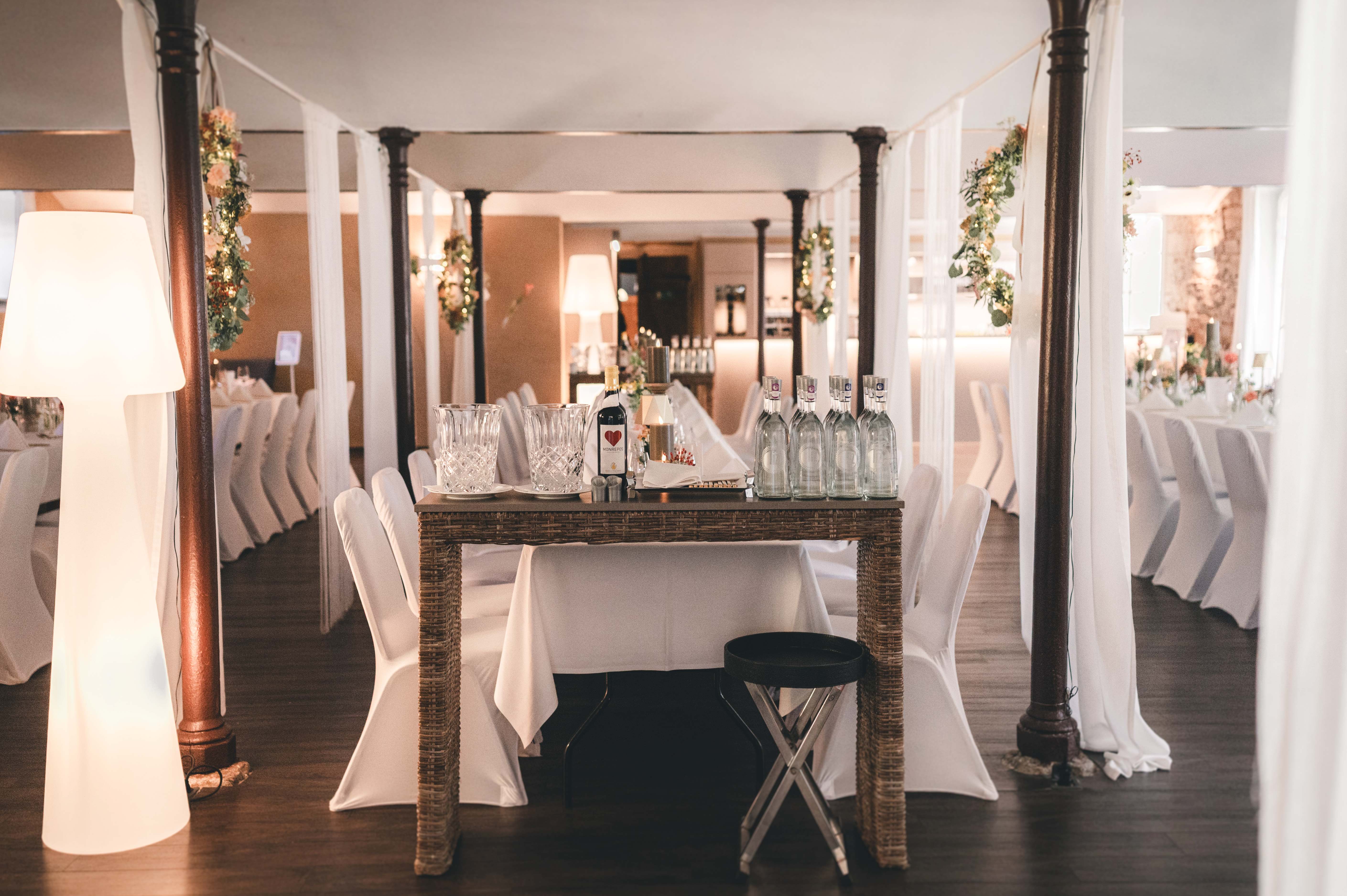 Innenansicht des elegant dekorierten Festsaals mit weißen Stuhlhussen, hängenden Blumenkränzen und festlich gedeckten langen Tafeln.