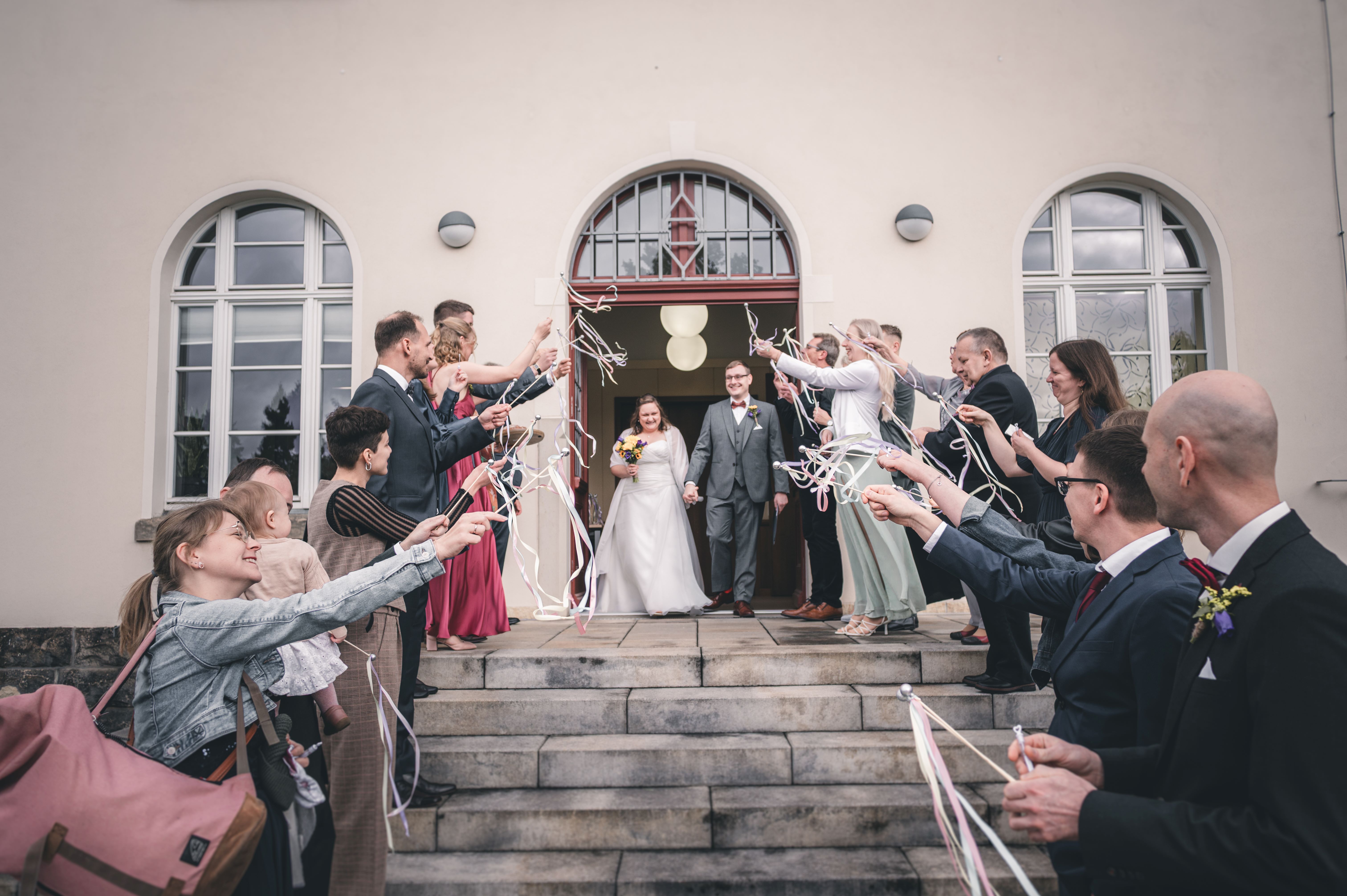 Das frischgebackene Ehepaar verlässt das Standesamt Radebeul durch ein fröhliches Spalier aus Gästen, die bunte Wedding Wands schwingen.