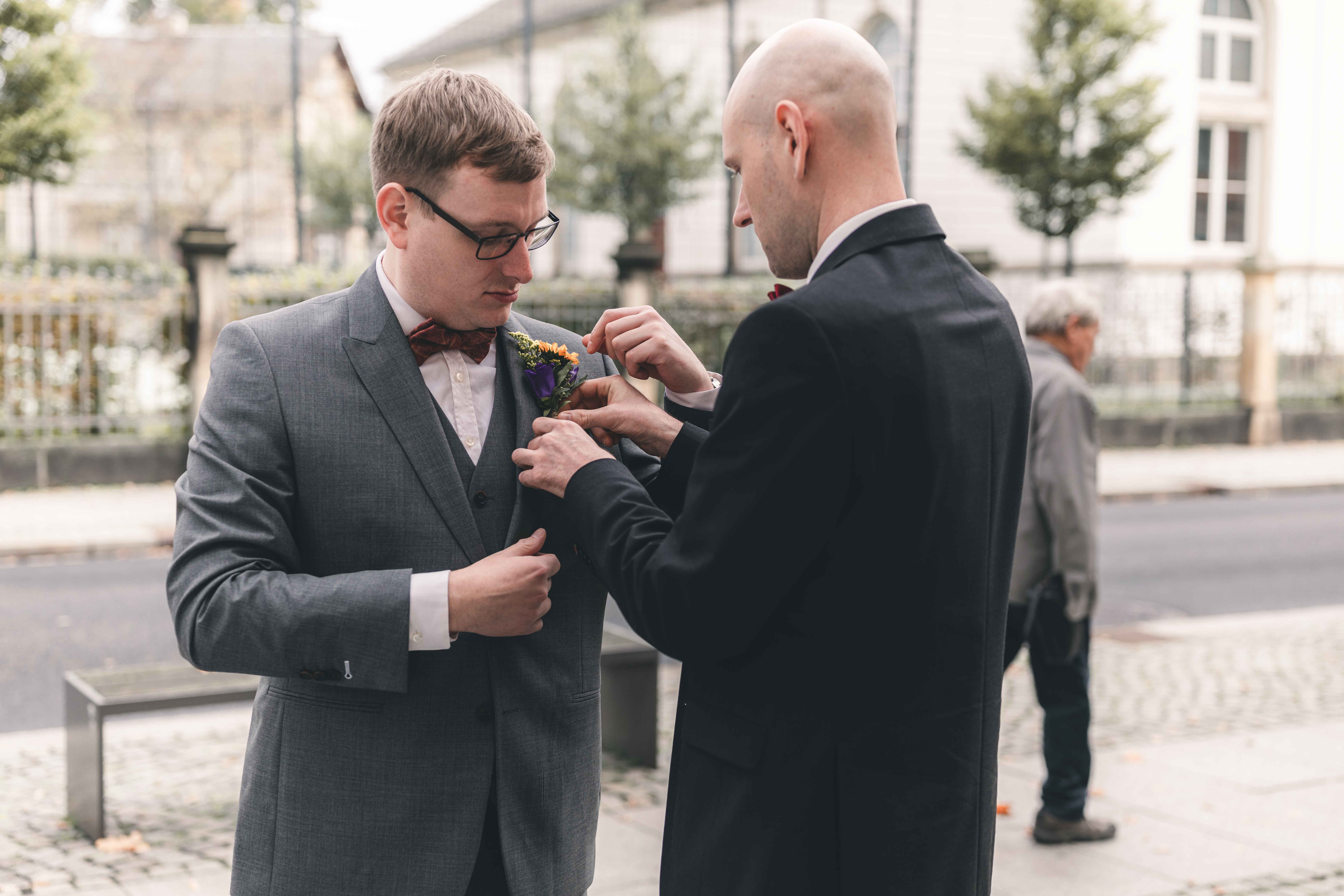 Der Trauzeuge bringt die bunte Ansteckblume (Boutonnière) am grauen Hochzeitsanzug des Bräutigams vor dem Standesamt Radebeul an.