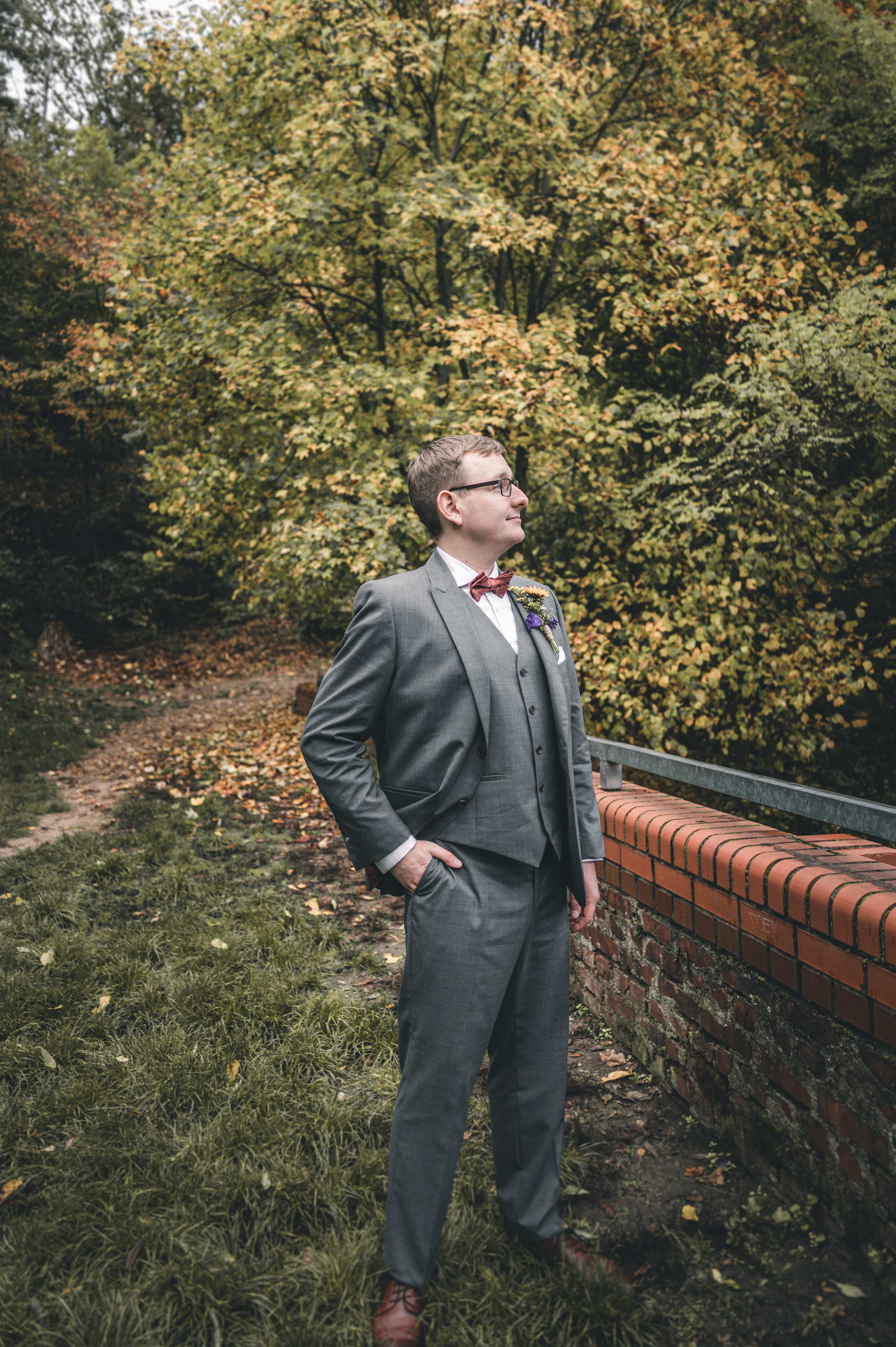 Der Bräutigam im eleganten grauen Dreiteiler steht mit einer Hand in der Tasche im Profil vor herbstlichen Bäumen und blickt entspannt zur Seite.