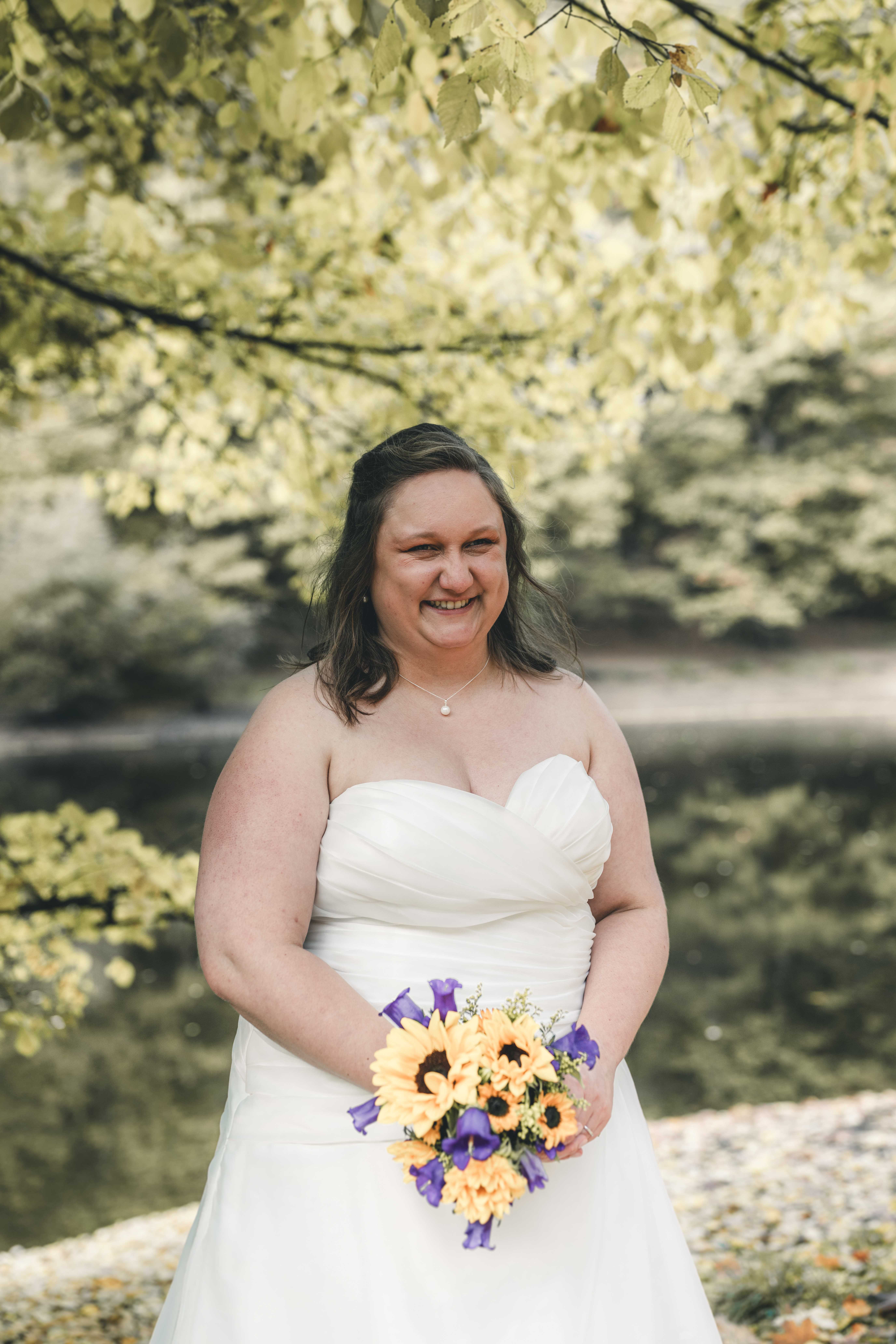 Ein wunderschönes Porträt der lächelnden Braut im schulterfreien Hochzeitskleid, die ihren gelb-violetten Sonnenblumenstrauß vor sich hält.
