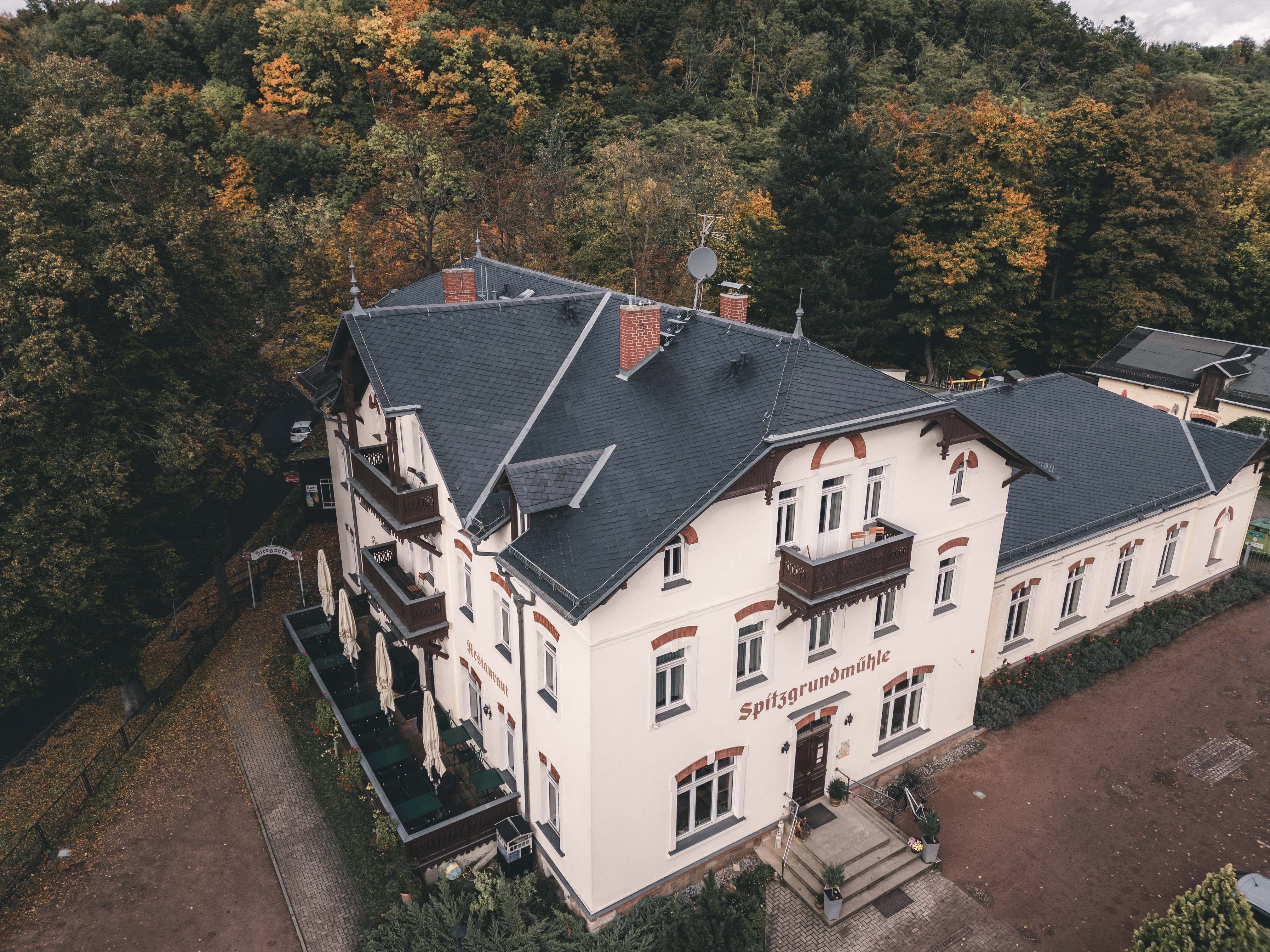 Eine beeindruckende Drohnen-Luftaufnahme der Spitzgrundmühle in Coswig bei Dresden, umgeben von dichten, herbstlich gefärbten Wäldern.