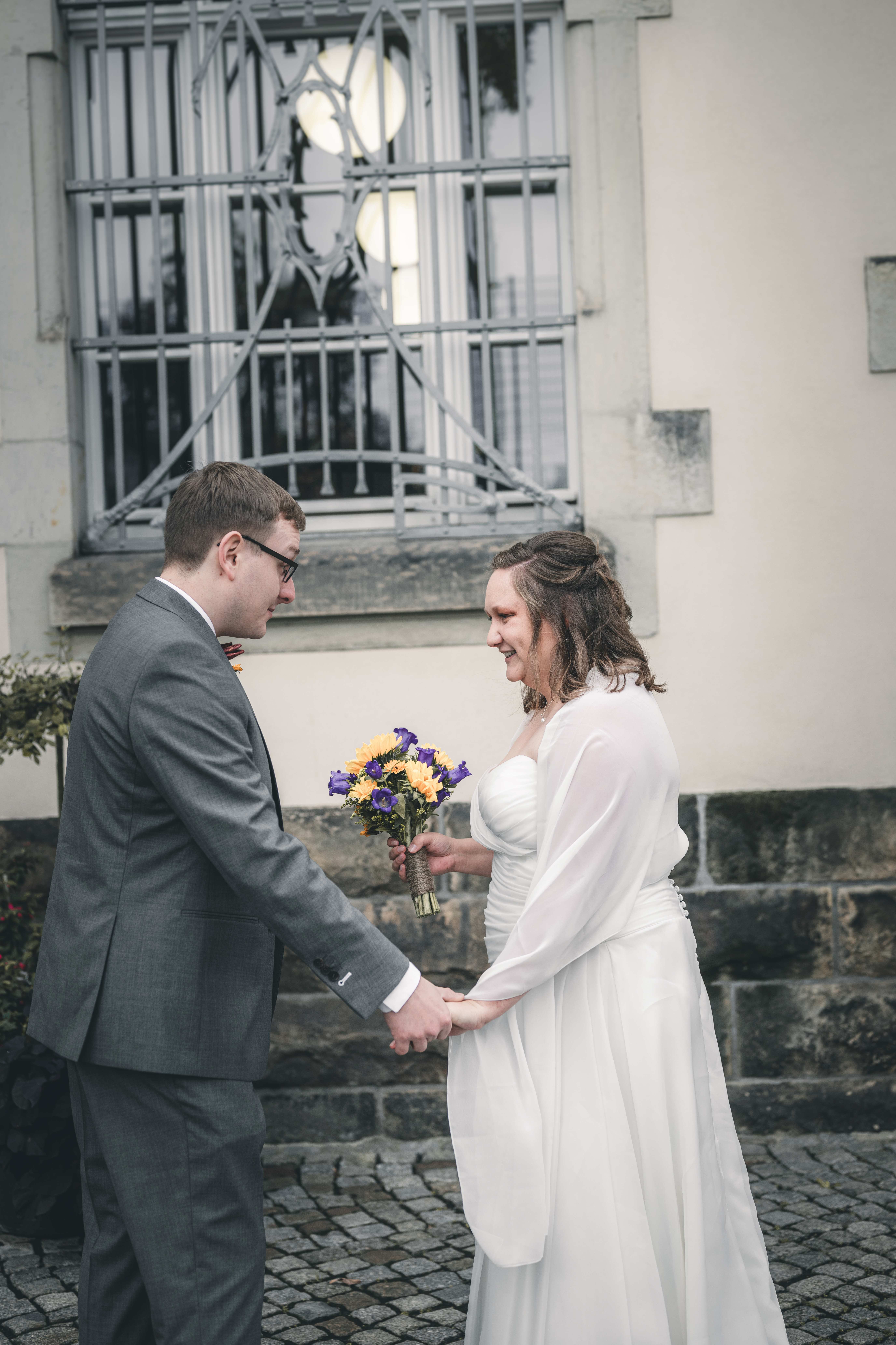 Das Brautpaar sieht sich beim First Look vor dem Standesamt Radebeul bei Dresden glücklich zum ersten Mal am Hochzeitstag, während die Braut einen gelb-violetten Strauß hält.