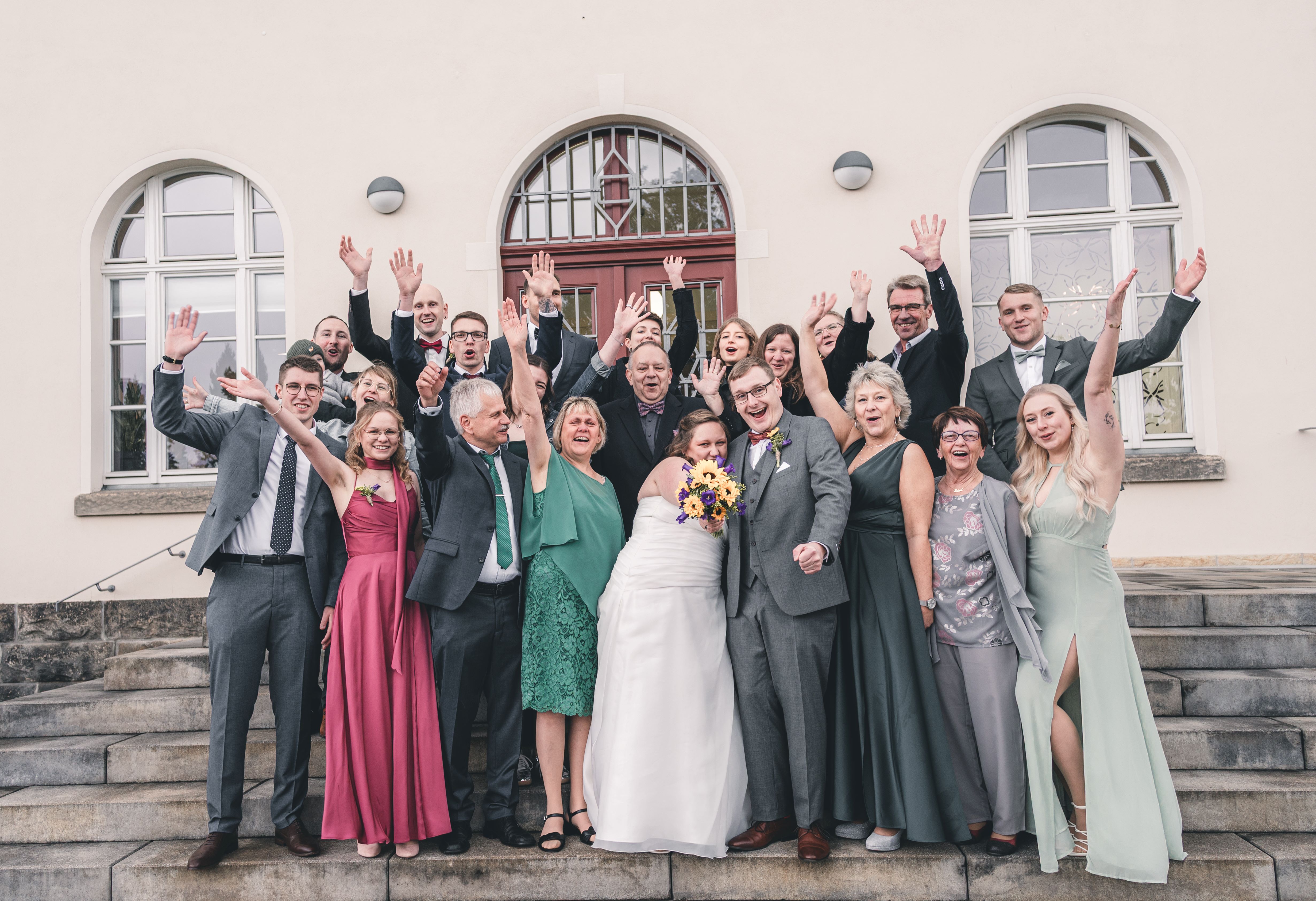 Die gesamte Hochzeitsgesellschaft posiert lachend mit erhobenen Händen für ein großes Gruppenbild auf den Steinstufen vor dem Standesamt Radebeul.