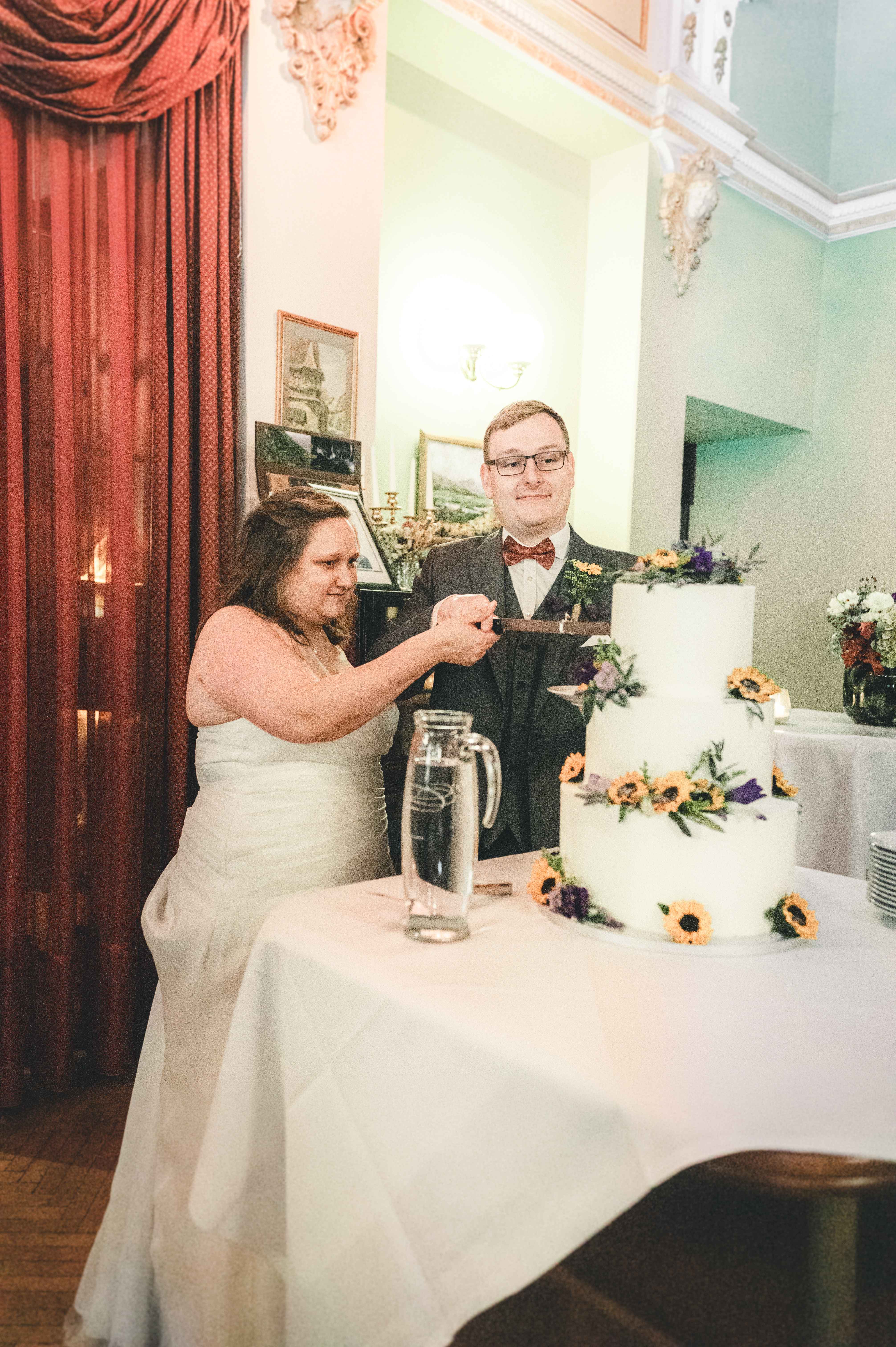 Das Brautpaar hält gemeinsam das Messer und schneidet lächelnd die dreistöckige Hochzeitstorte mit echten Sonnenblumen im Festsaal in Coswig an.