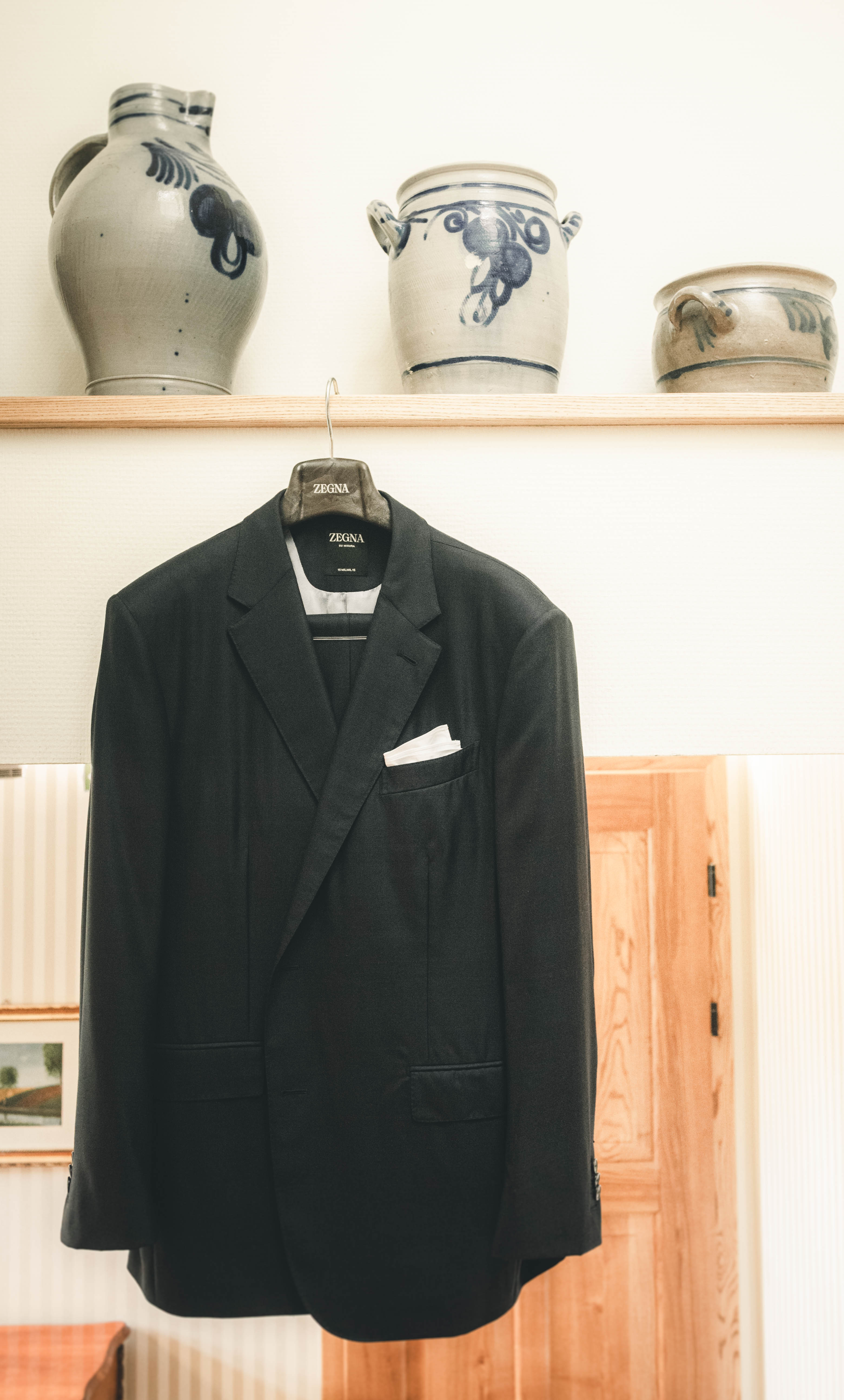 Ein dunkler Hochzeitsanzug von Zegna mit weißem Einstecktuch hängt an einem Holzregal unter traditionellen Steingutkrügen im Magdalenengut Erfurt.