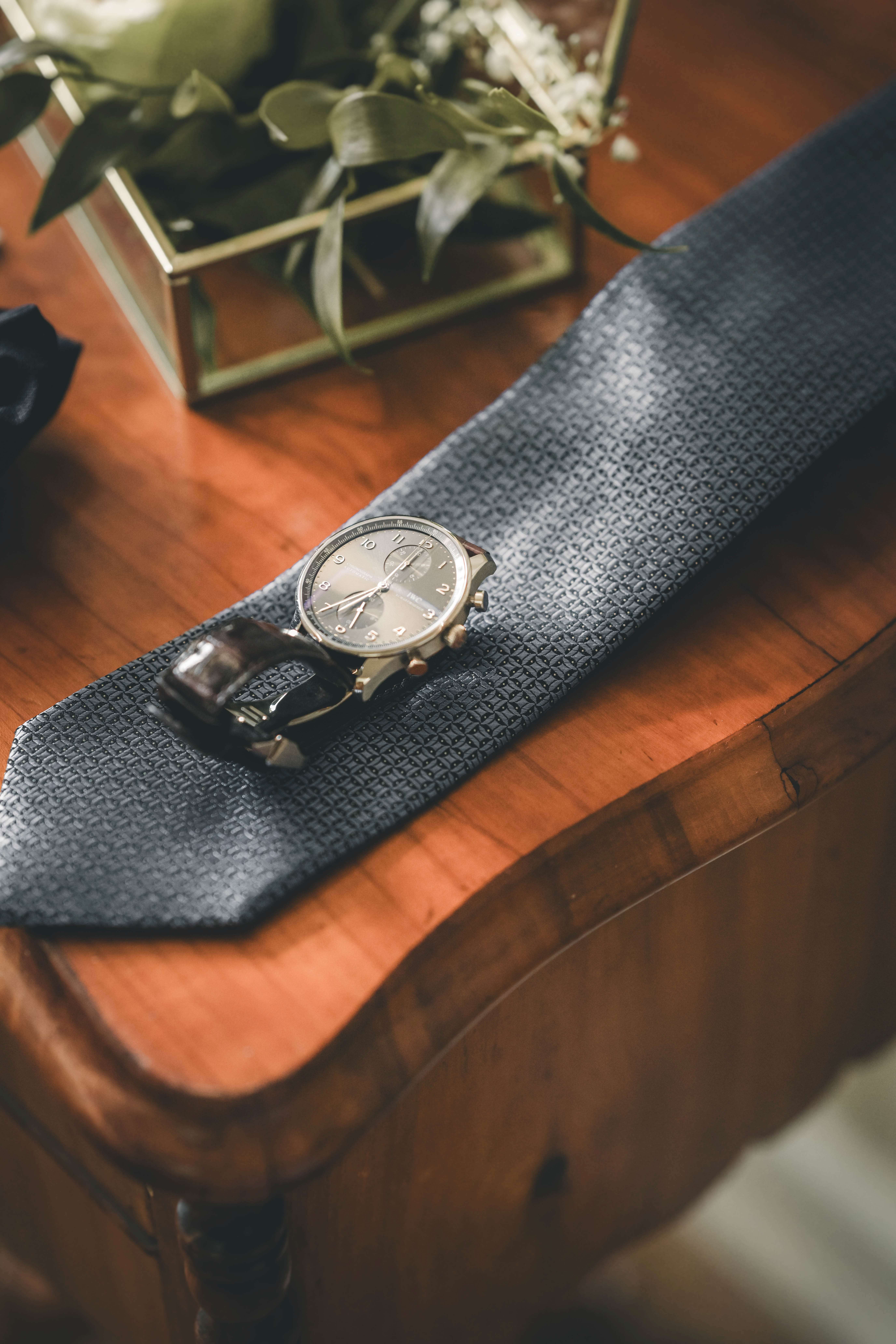 Detailaufnahme einer IWC-Armbanduhr auf einer dunkelblauen gemusterten Hochzeitskrawatte auf poliertem Holz.