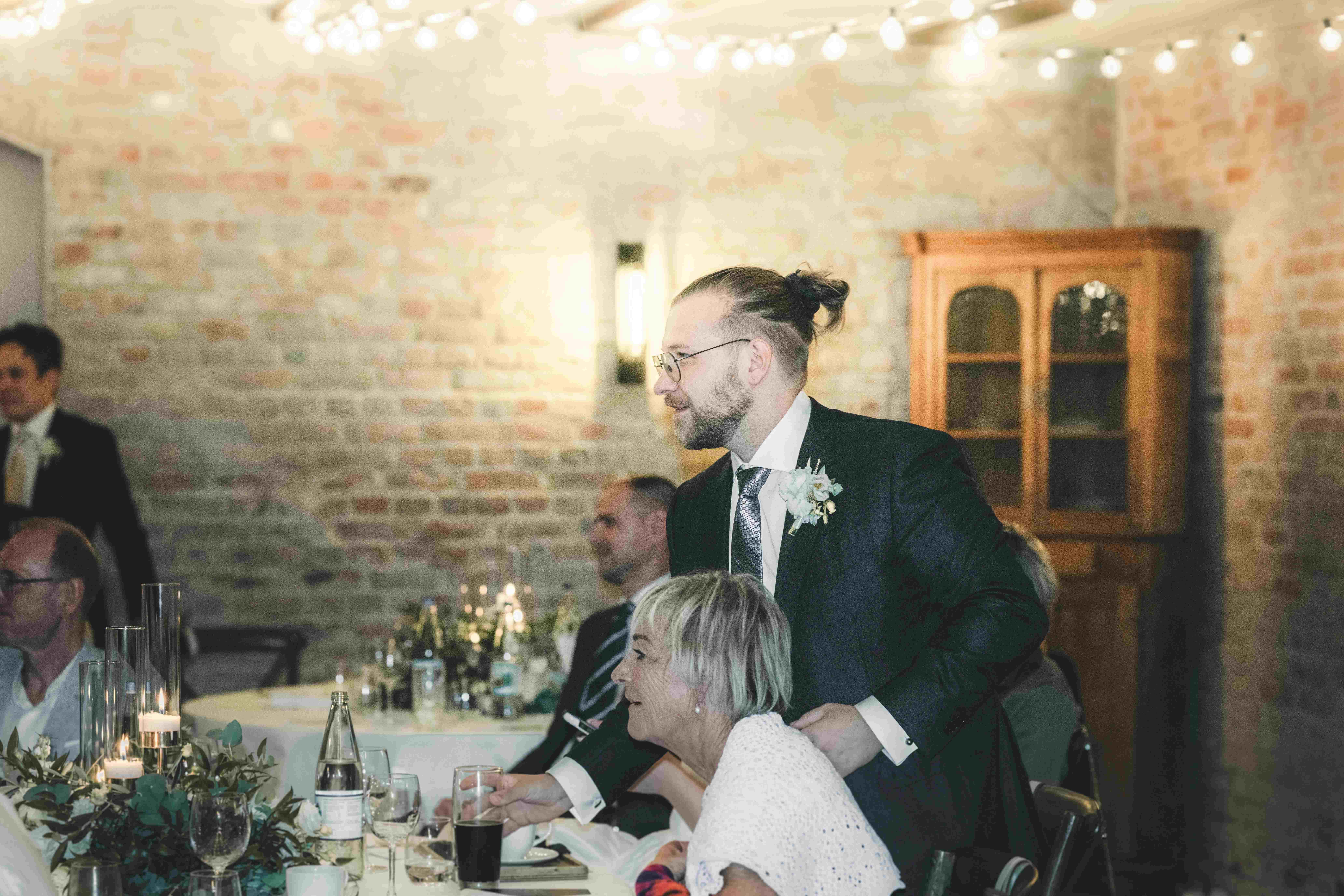 Der Bräutigam mit Brille und Ansteckblume steht lächelnd an der festlich dekorierten Hochzeitstafel im Saal des Magdalenenguts Erfurt.