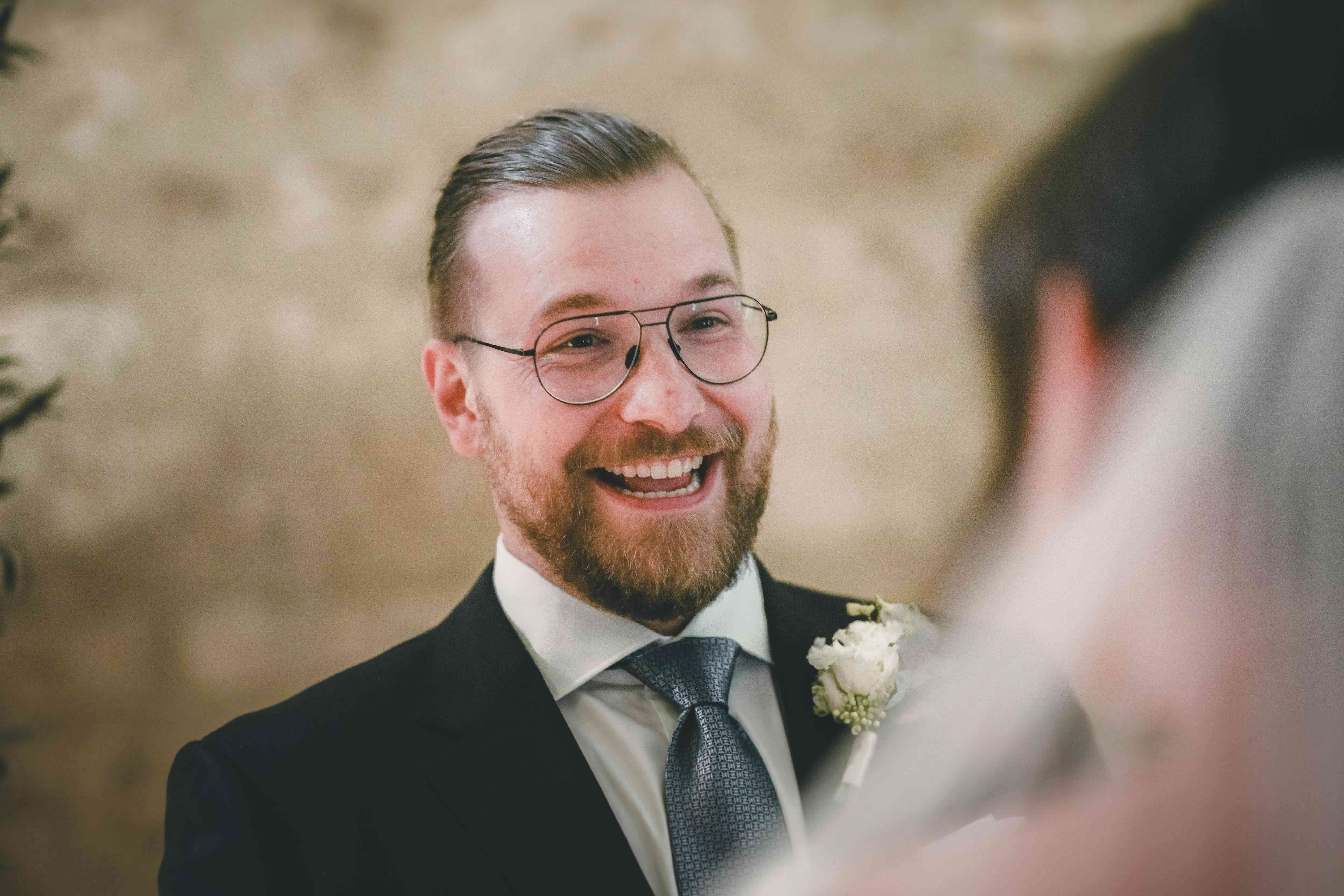 Nahaufnahme des Bräutigams mit Brille, Krawatte und Boutonnière, der seine Braut während der Trauzeremonie im Magdalenengut Erfurt glücklich anstrahlt.