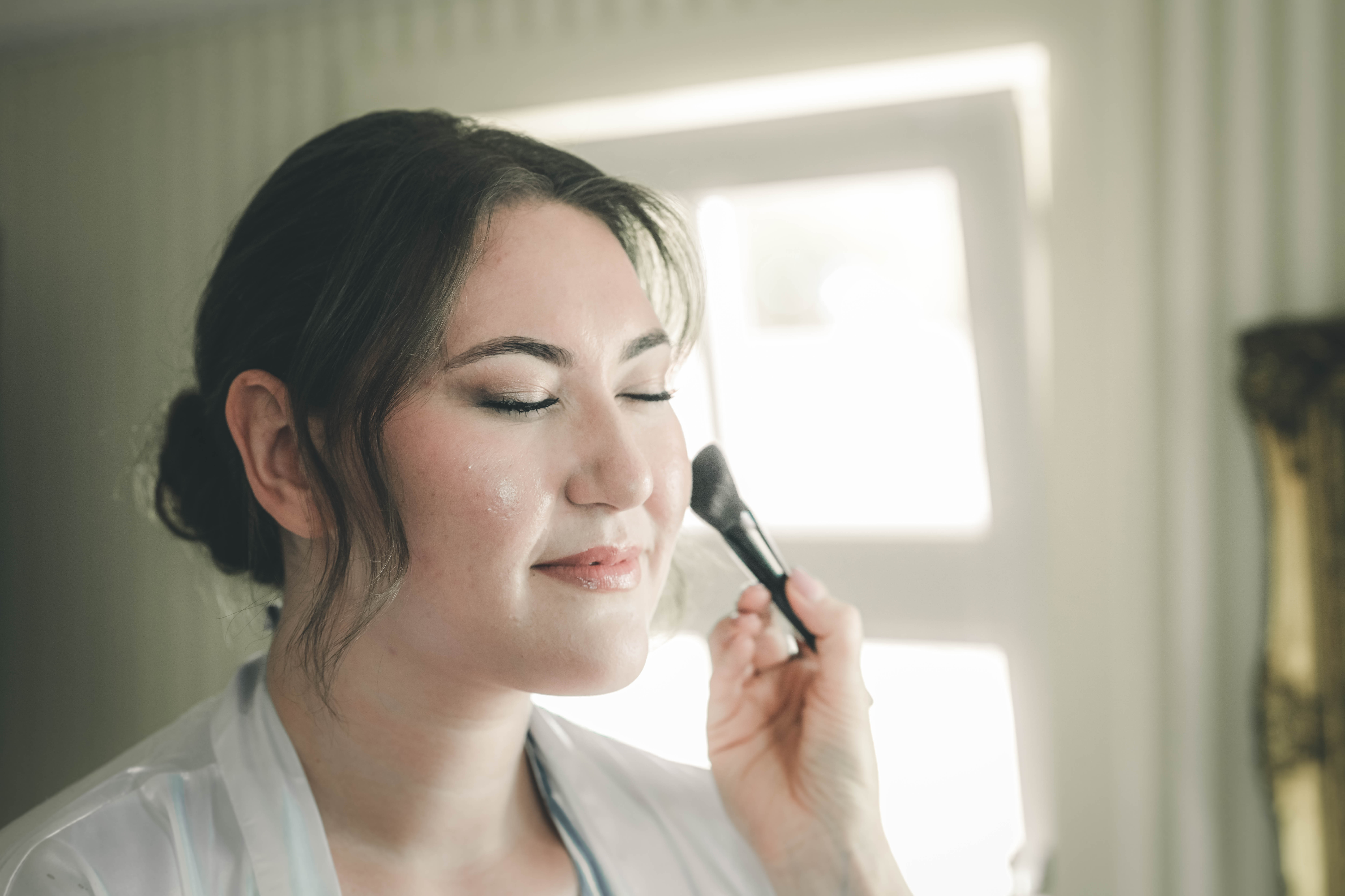 Nahaufnahme der Braut mit geschlossenen Augen und sanftem Lächeln, während die Stylistin mit einem weichen Pinsel Puder aufträgt.