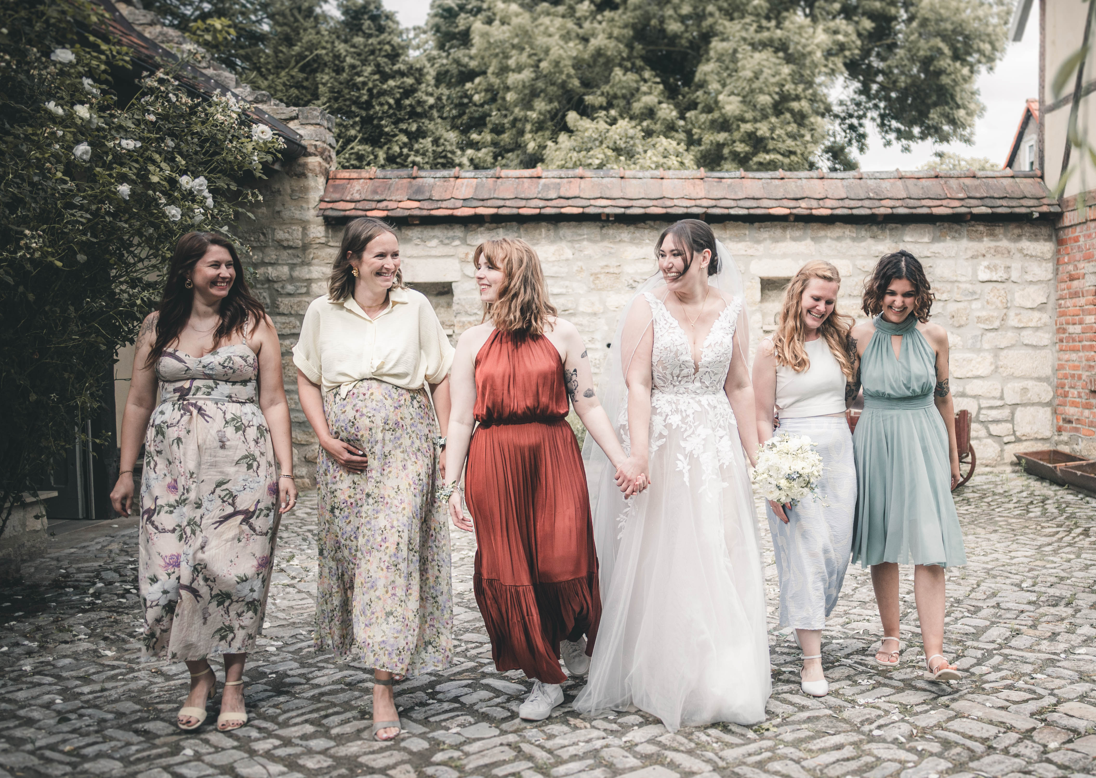Die Braut im weißen Spitzenkleid schlendert lachend mit ihren Freundinnen und Brautjungfern über den Hof des Magdalenenguts Erfurt.
