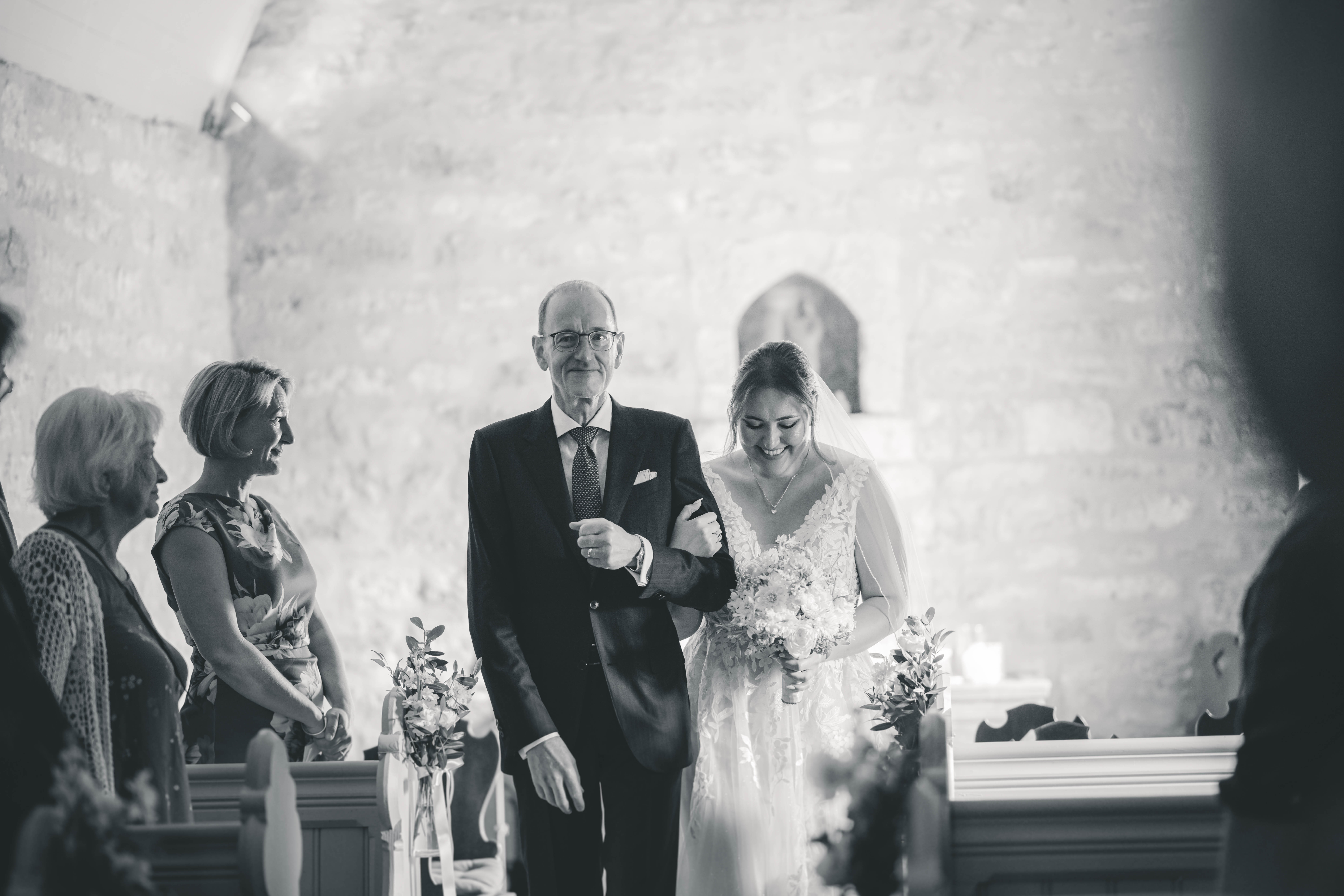 Schwarz-Weiß-Fotografie: Die lächelnde Braut blickt gerührt nach unten, während sie mit ihrem Vater an den Kirchenbänken und Gästen im Magdalenengut Erfurt vorbeigeht.
