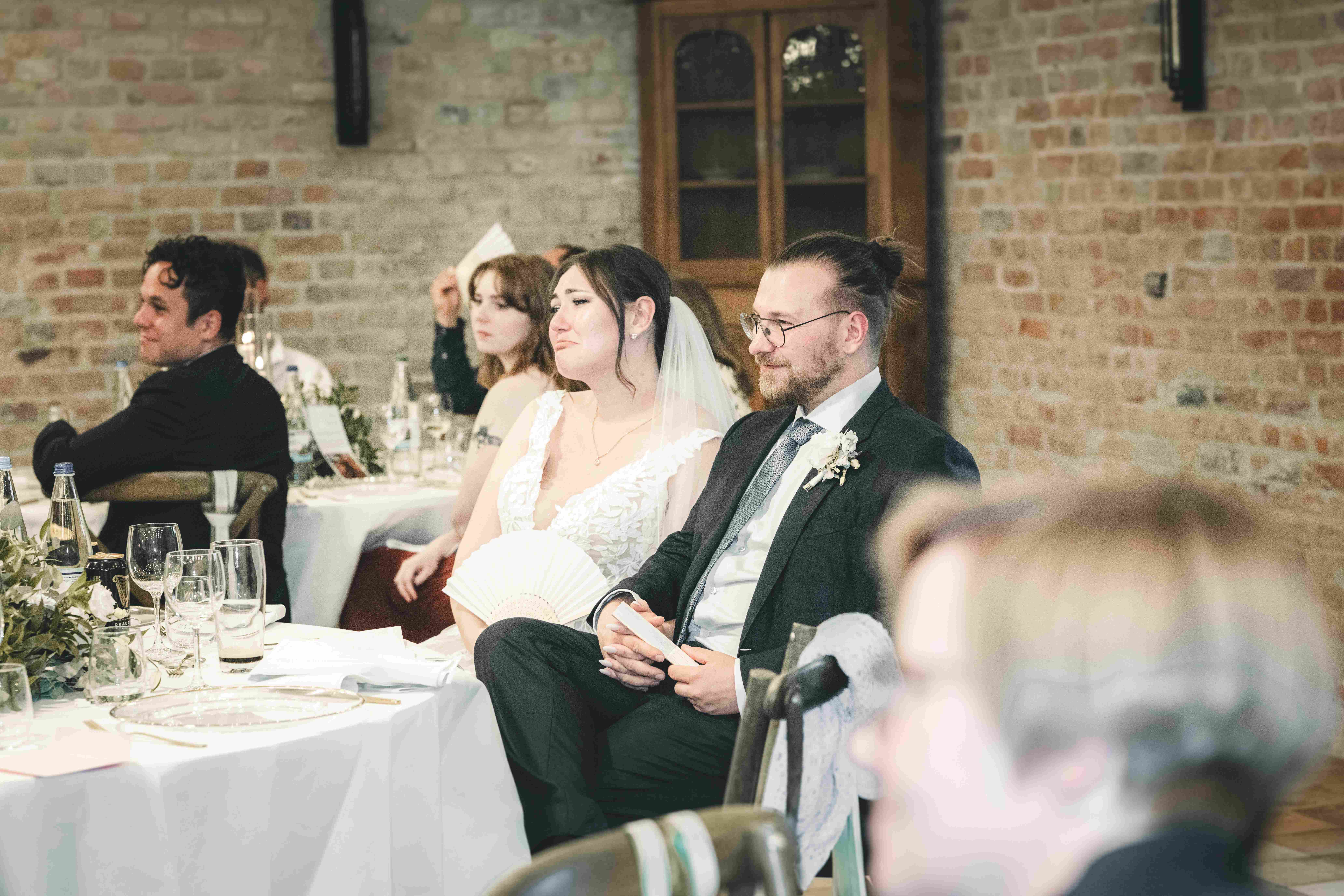 Die Braut wischt sich mit Fächer in der Hand Freudentränen ab, während sie mit dem Bräutigam am Dinnertisch im Magdalenengut Erfurt sitzt.