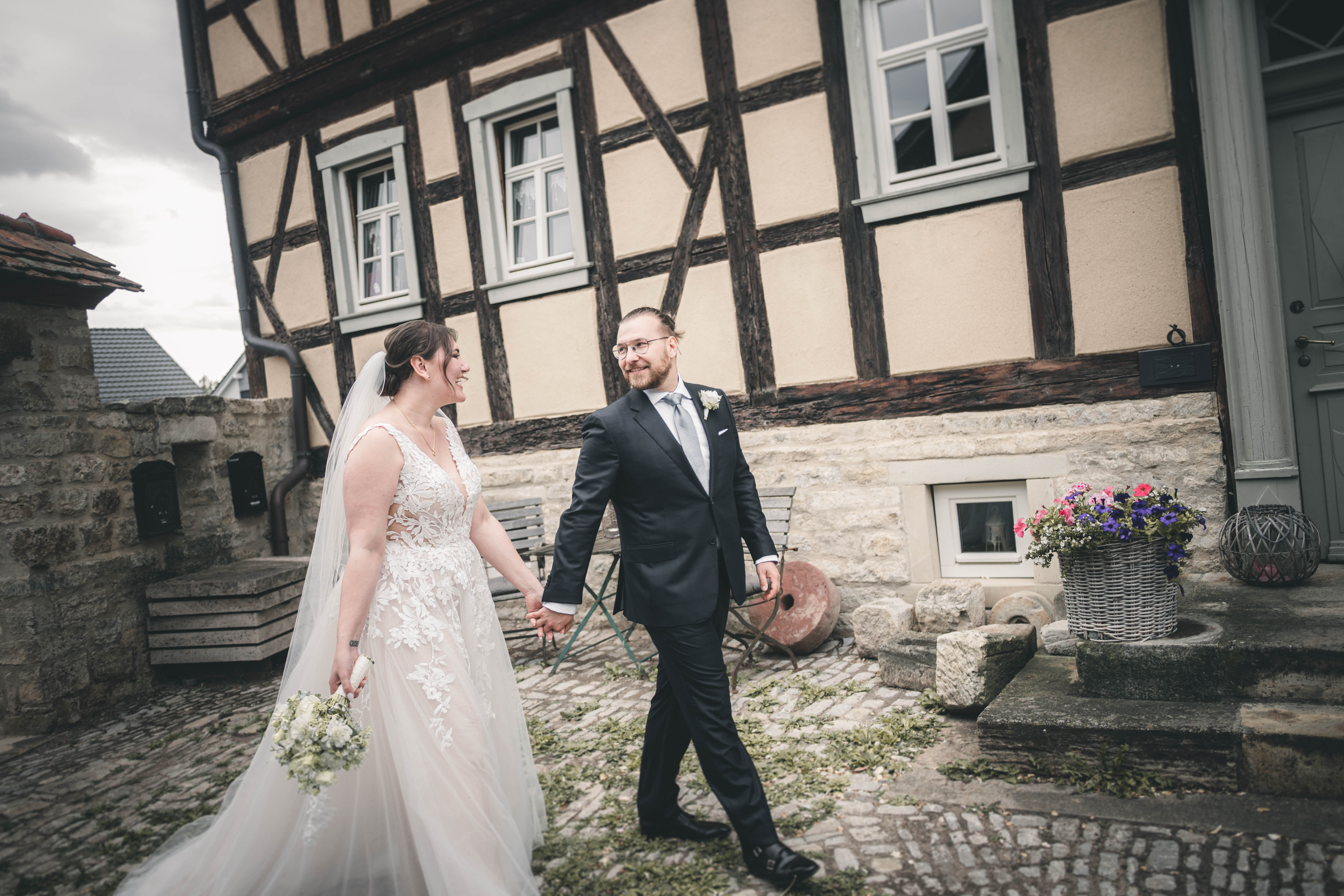 Braut und Bräutigam spazieren Hand in Hand über den Hof und blicken sich vor dem großen Fachwerkgebäude des Magdalenenguts Erfurt lächelnd an.