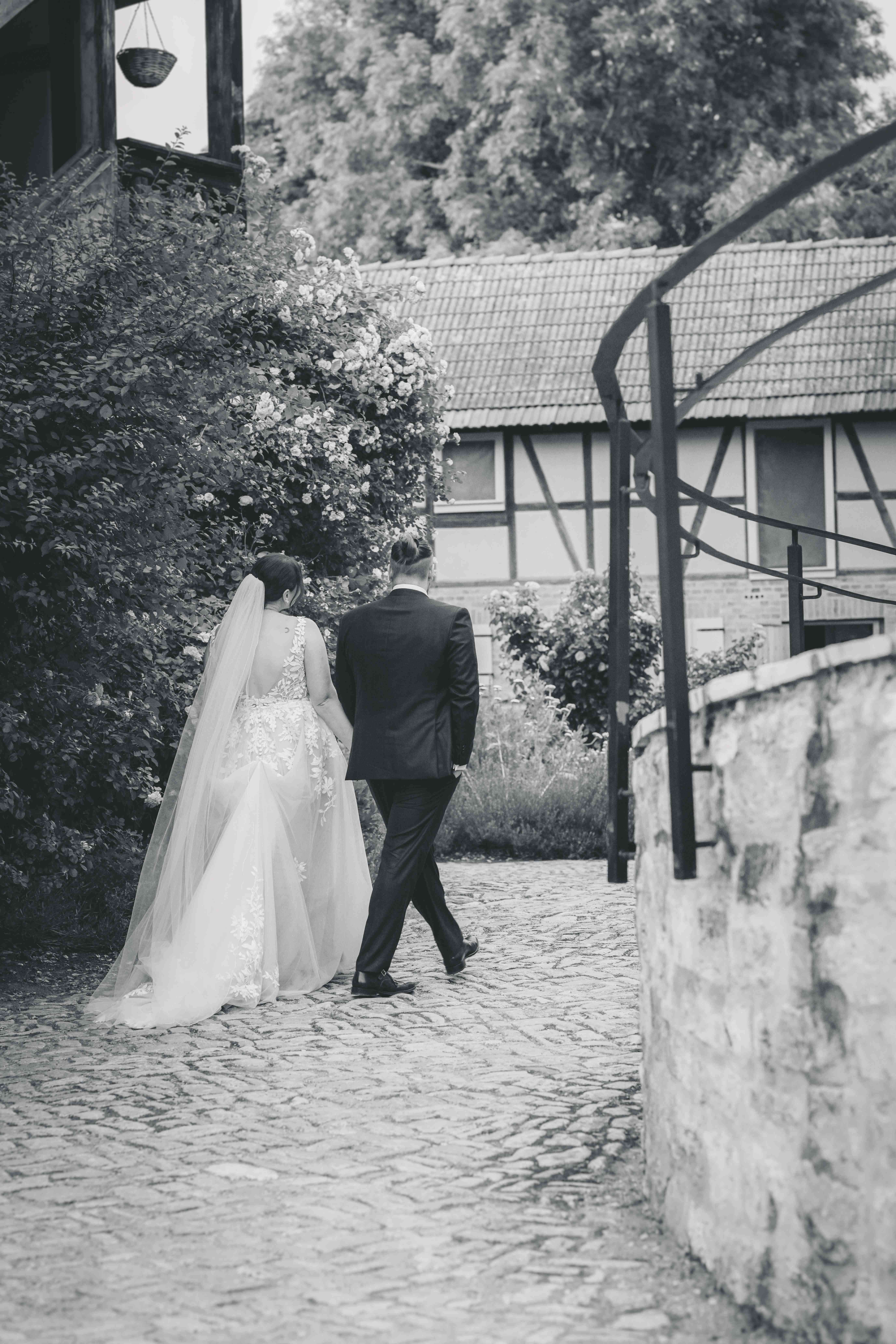 Schwarz-Weiß-Fotografie des Brautpaars von hinten, wie sie über das Pflaster am Fachwerkhaus im Magdalenengut Erfurt entlanggehen.