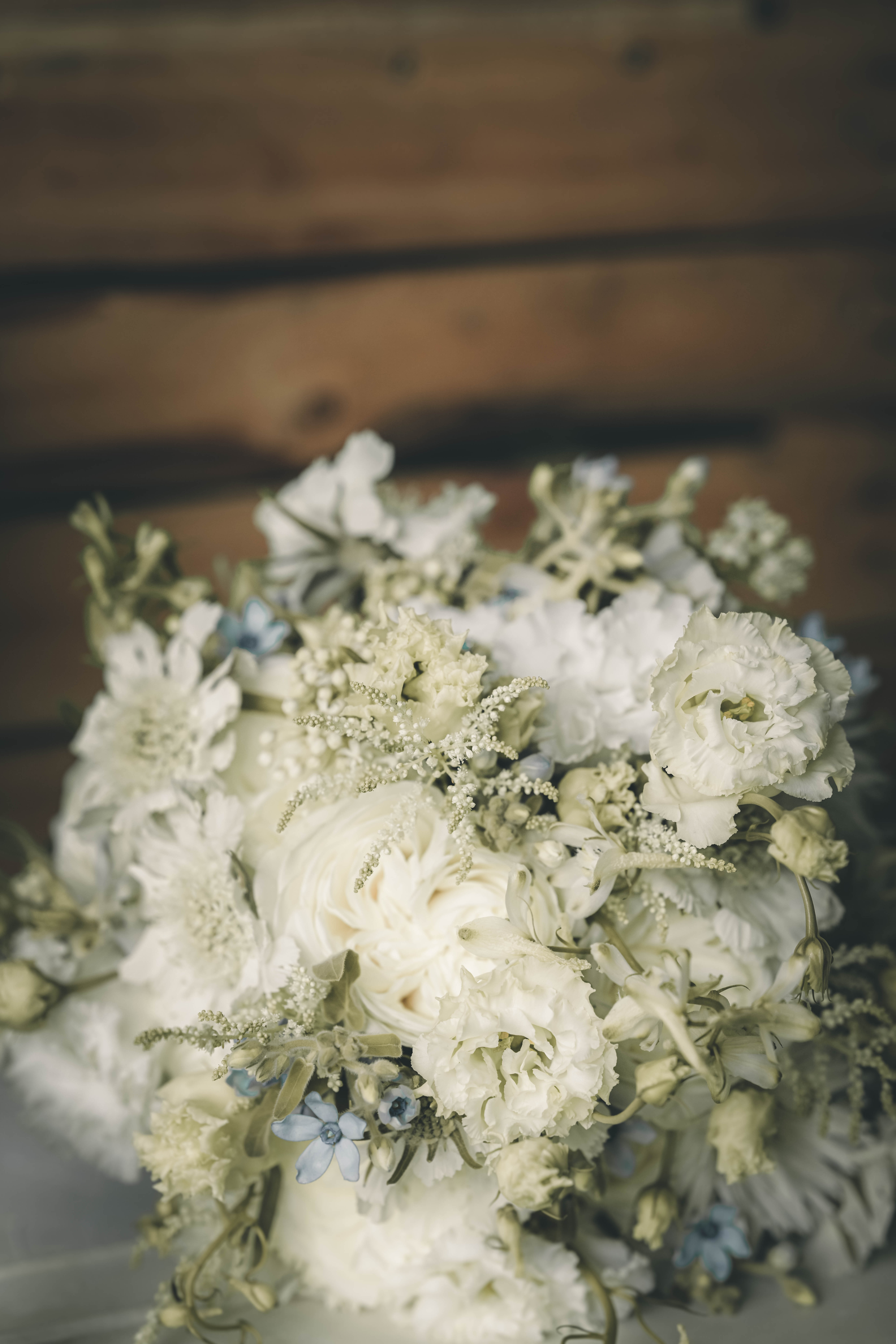 Detailaufnahme eines edlen weißen Hochzeitsstraußes aus Rosen, Lisianthus und zartblauen Akzenten vor einer rustikalen Holzwand im Magdalenengut Erfurt.