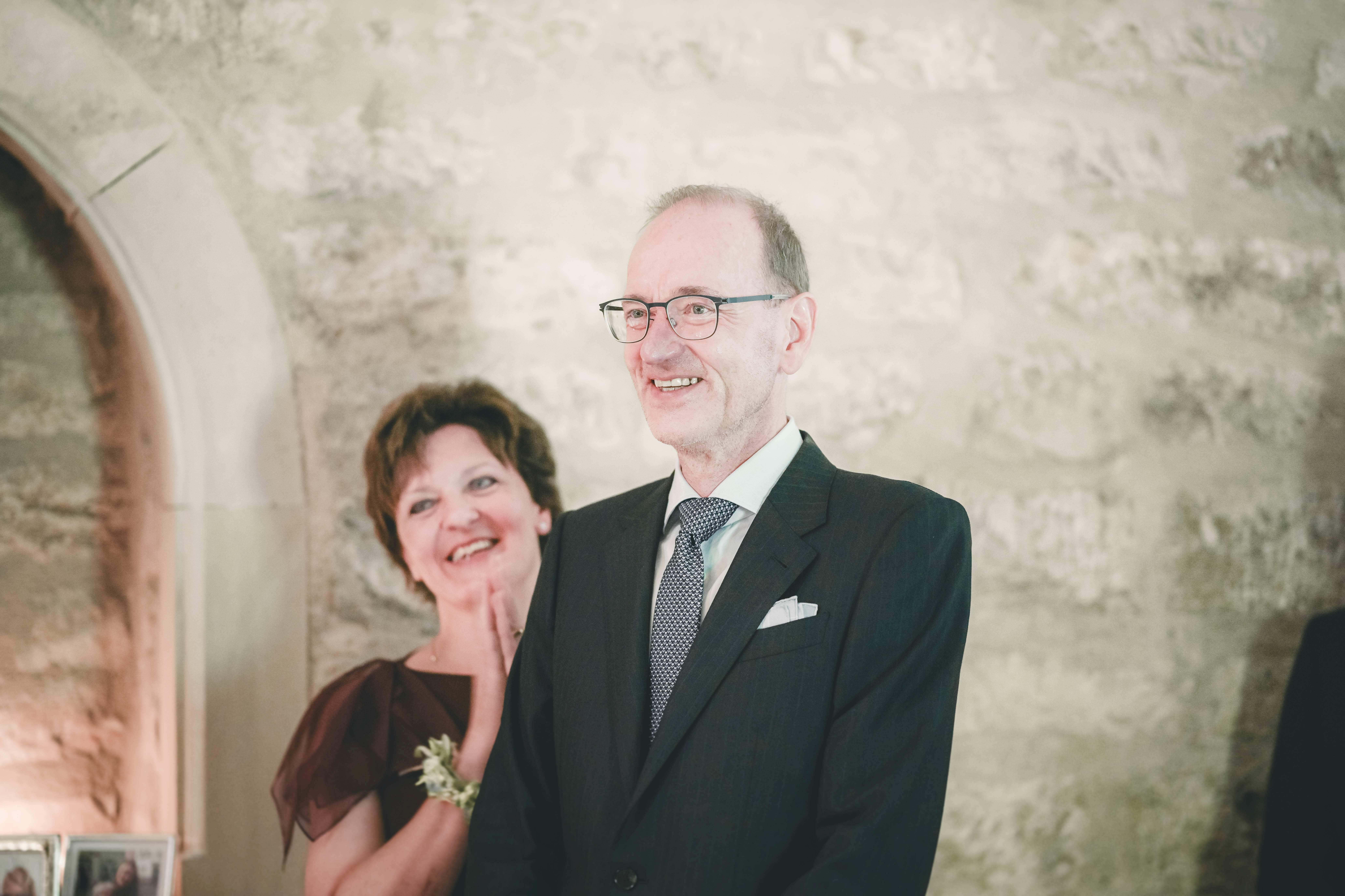 Der Brautvater im eleganten Anzug lächelt glücklich, während die Mutter im Hintergrund die Hände vor Freude zusammenlegt.