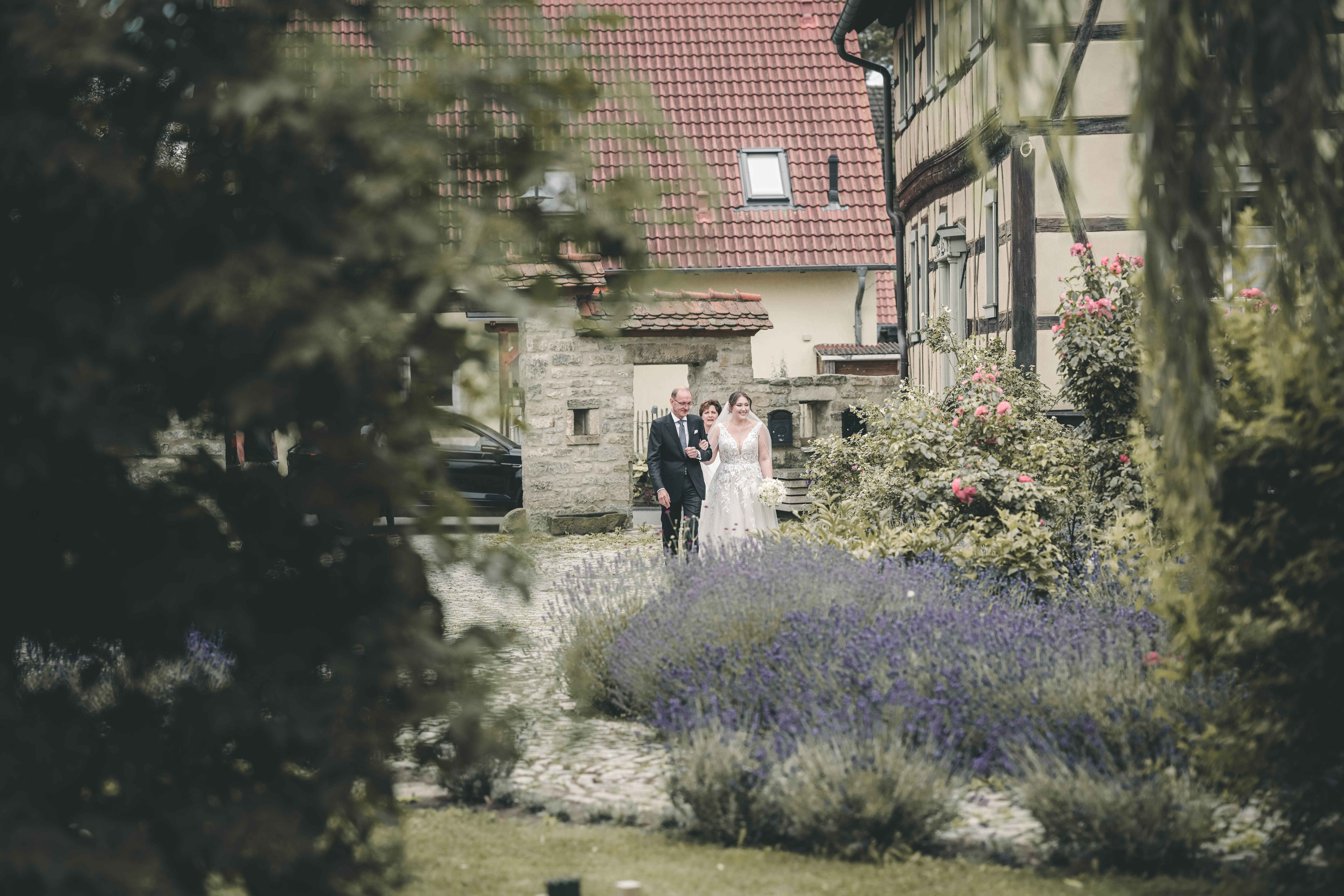 Die Braut im weißen Brautkleid geht in Begleitung ihrer Eltern über den gepflasterten Weg vorbei an blühendem Lavendel im Innenhof des Magdalenenguts Erfurt.