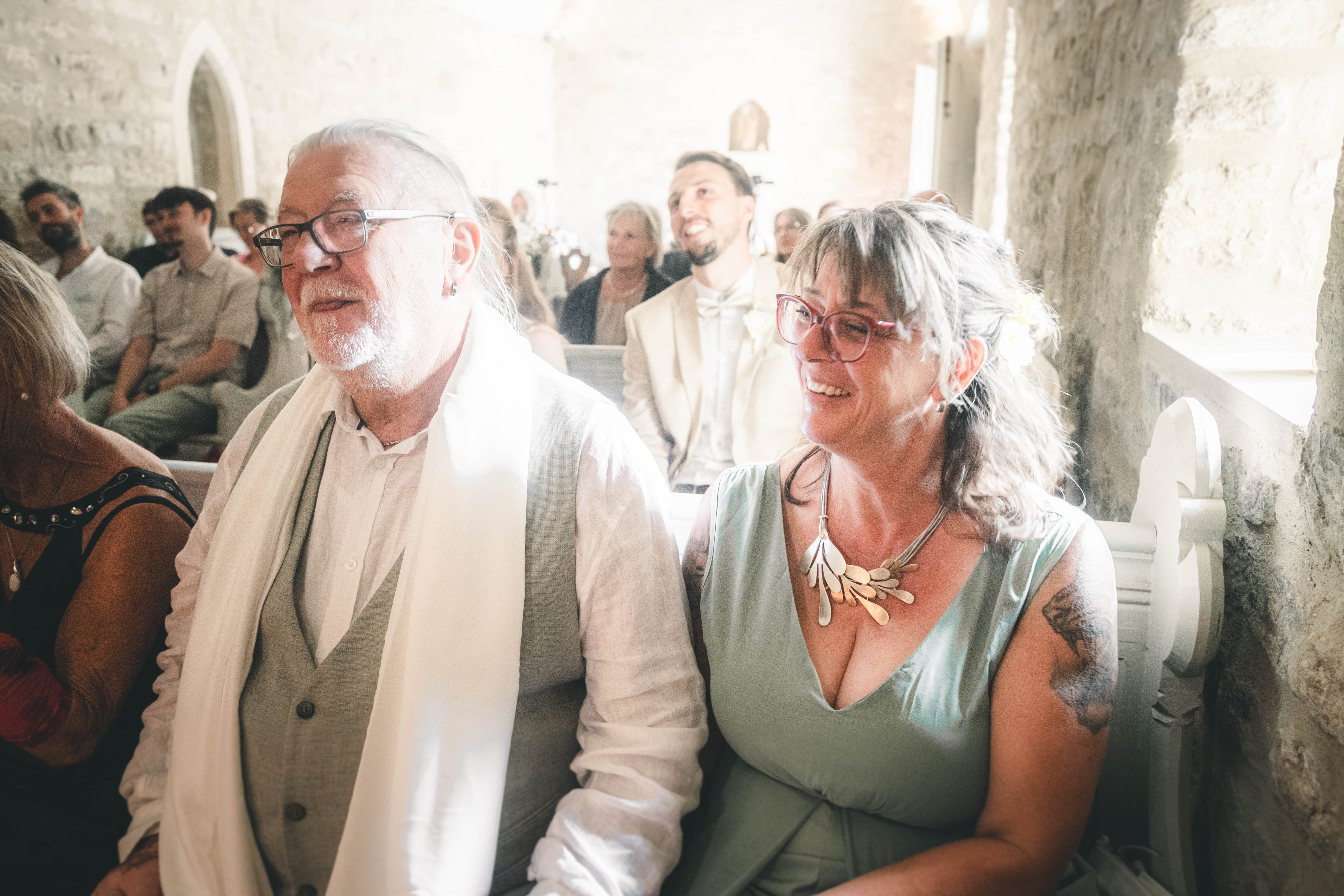 Ein älterer Herr mit weißem Schal und eine lächelnde Dame mit Brille und Kette verfolgen glücklich die Trauung in der ersten Reihe.