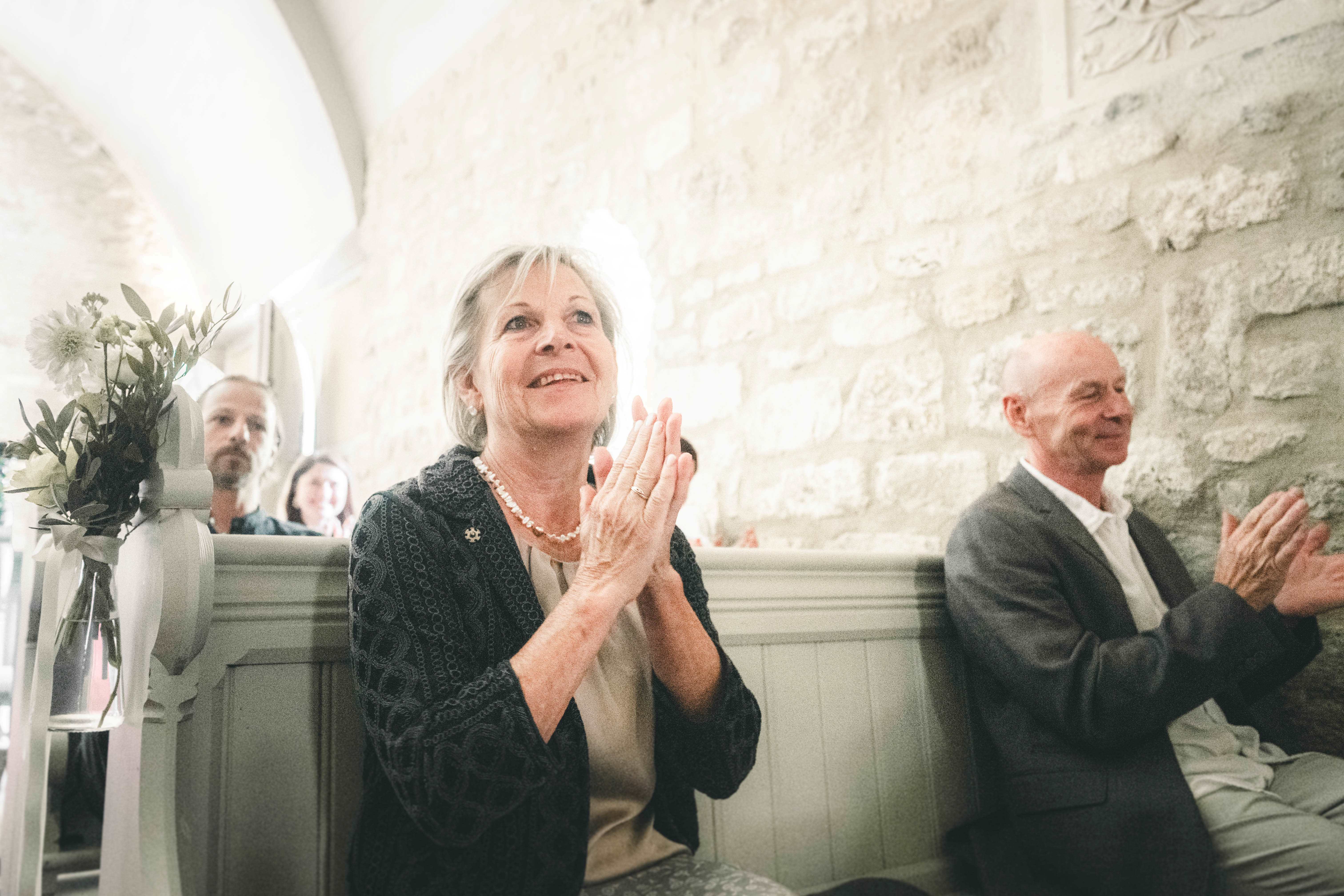 Eine strahlende Dame mit Perlenkette und ein Herr im Anzug applaudieren glücklich in der Kirchenbank der Kapelle im Magdalenengut Erfurt.