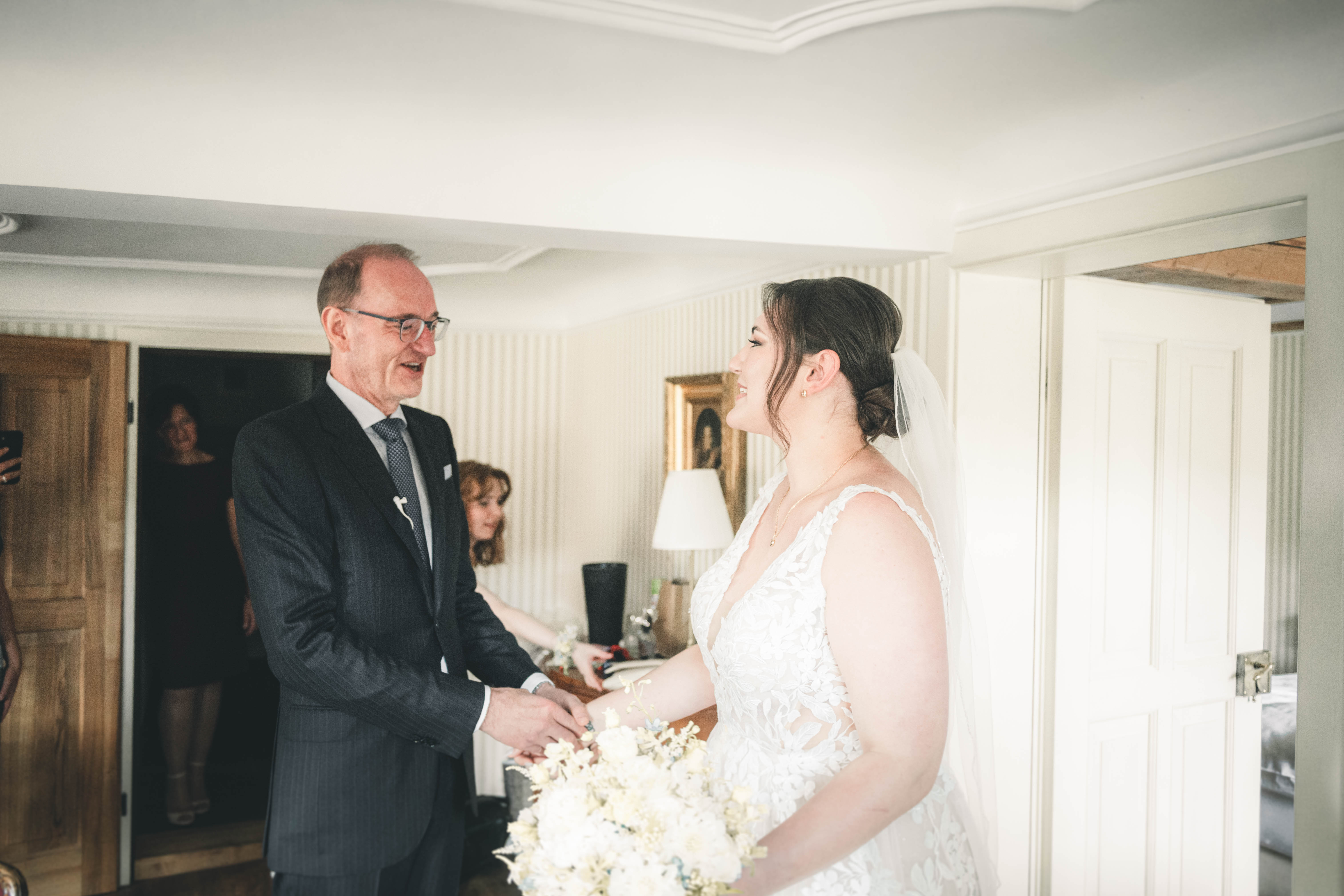 Der Brautvater im dunklen Anzug hält die Hände seiner strahlenden Tochter im Spitzenbrautkleid während des First Look im Magdalenengut Erfurt.