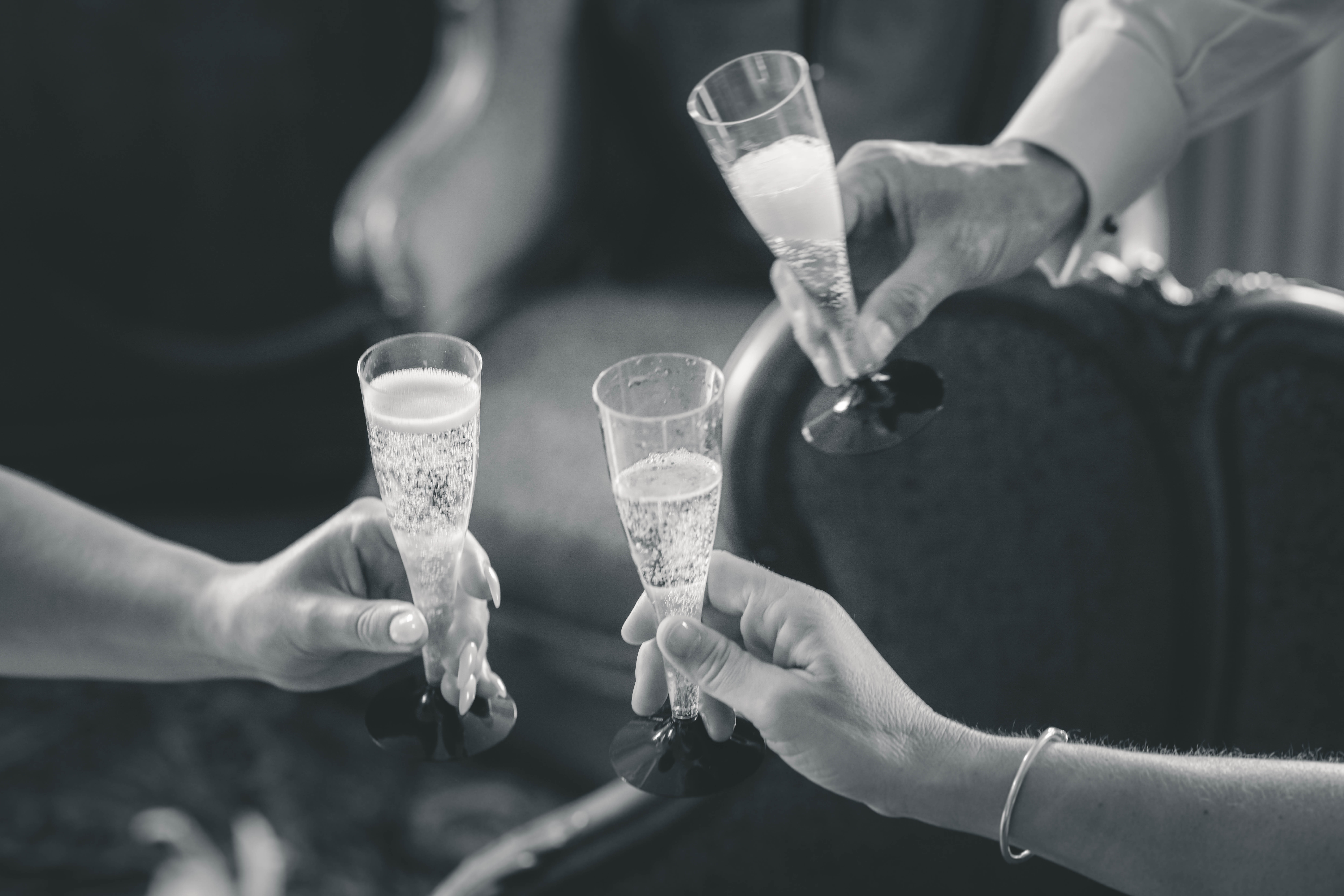 Drei Sektgläser, gefüllt mit Champagner, stoßen in Schwarz-Weiß an. Die Hände der anstoßenden Personen sind zu sehen.