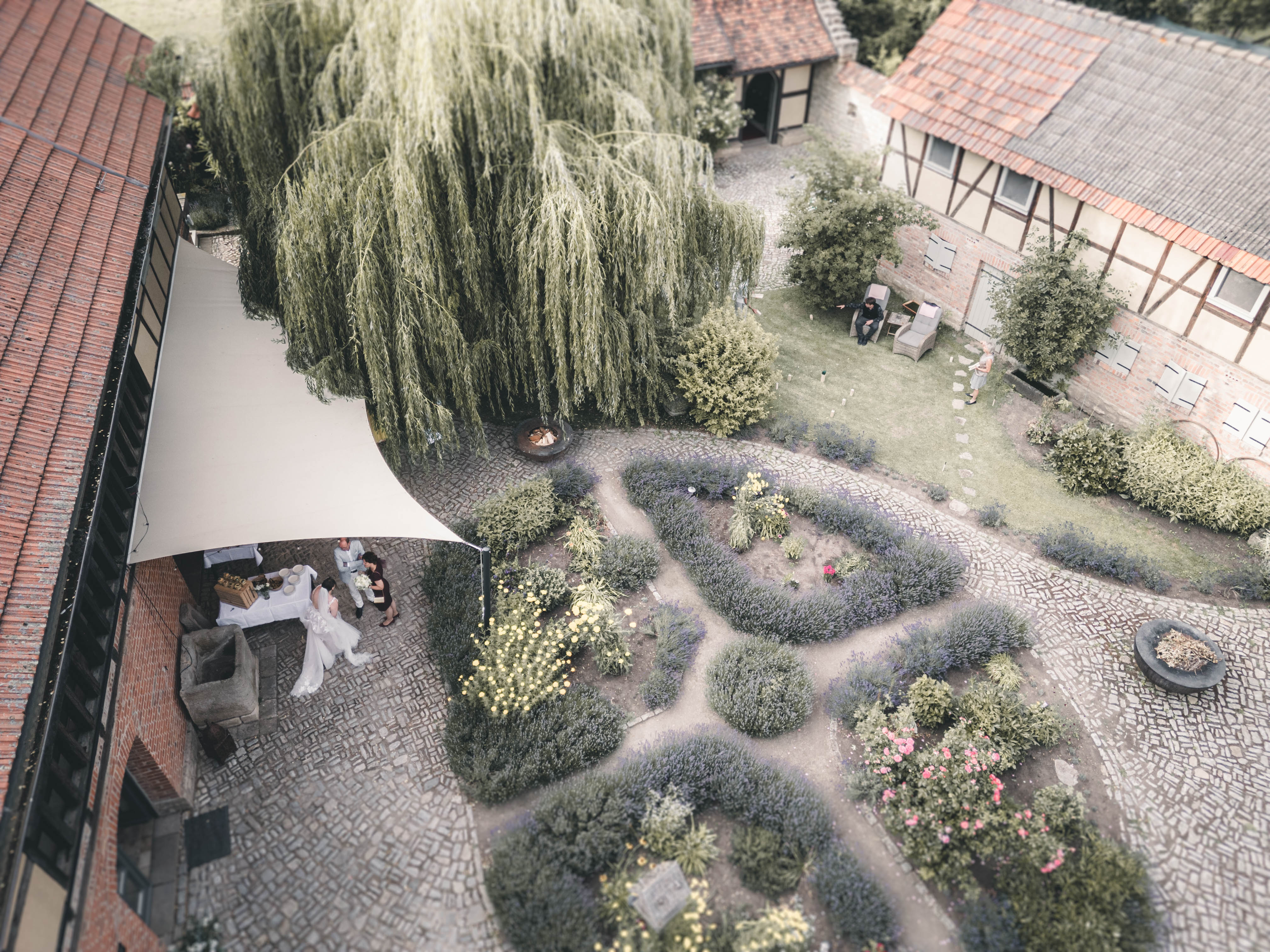Eine Luftaufnahme zeigt den malerischen Innenhof einer historischen Hochzeitslocation. Ein Brautpaar steht unter einem weißen Sonnensegel, umgeben von gepflegten Buchsbaumhecken, Lavendelbeeten und Rosen. Ein großer Weidenbaum spendet Schatten.