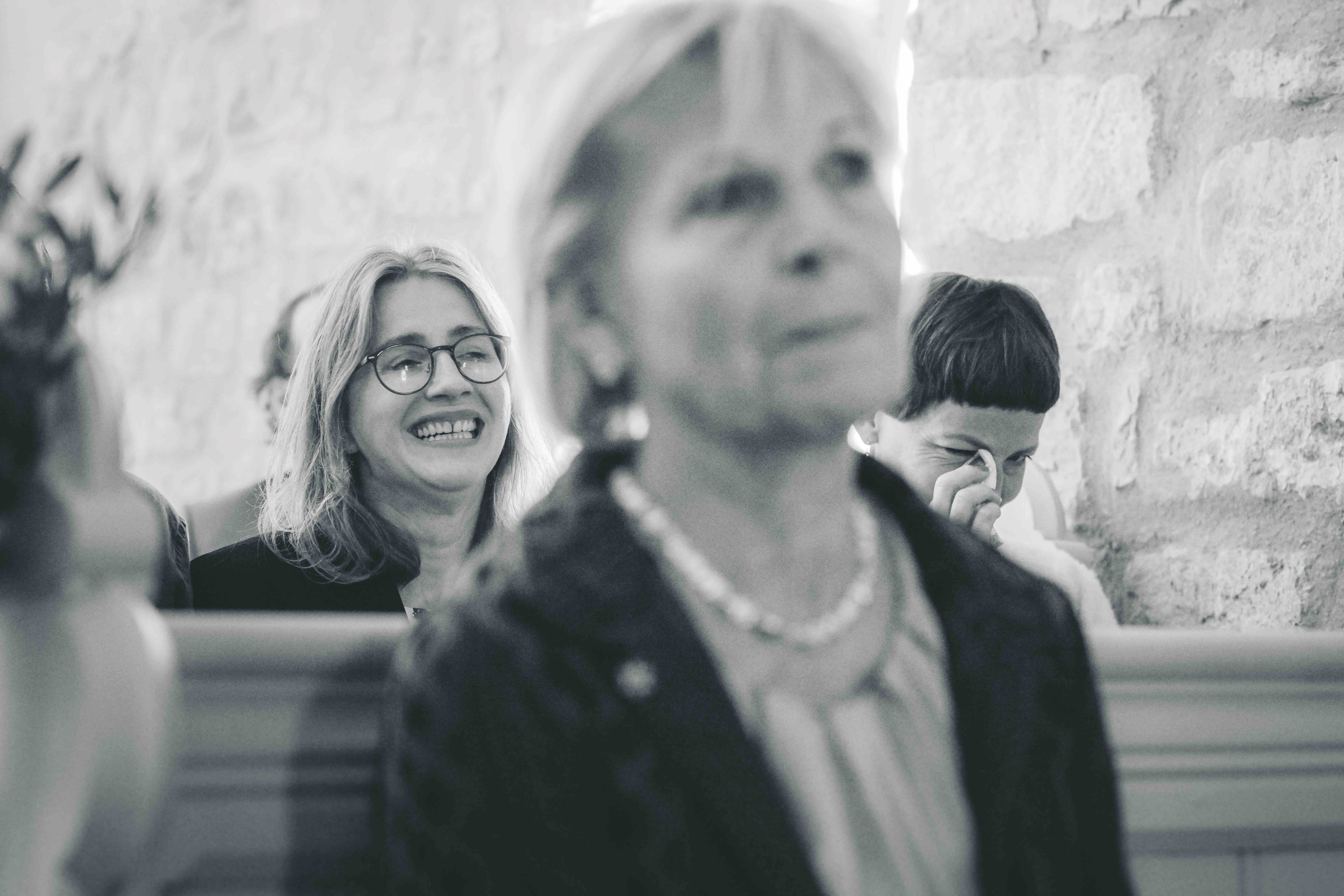 Schwarz-Weiß-Fotografie einer lachenden Frau mit Brille in der Kirchenbank, während ein Gast dahinter gerührt eine Freudenträne abtupft.