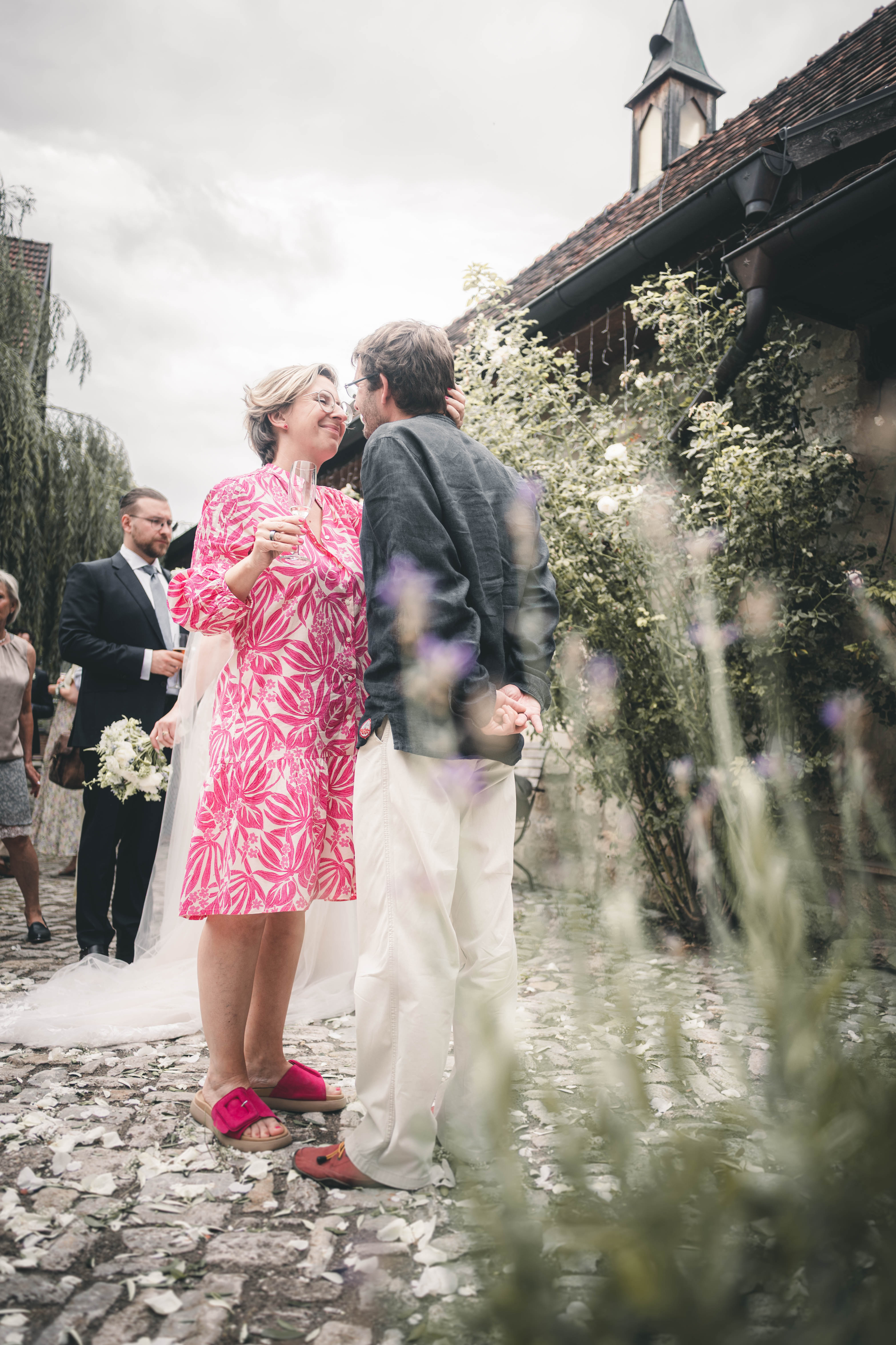 Ein Paar im gemusterten Kleid und dunklen Leinenhemd steht im innigen Dialog vor dem großen Rosenstrauch und der Kapelle des Magdalenenguts Erfurt.