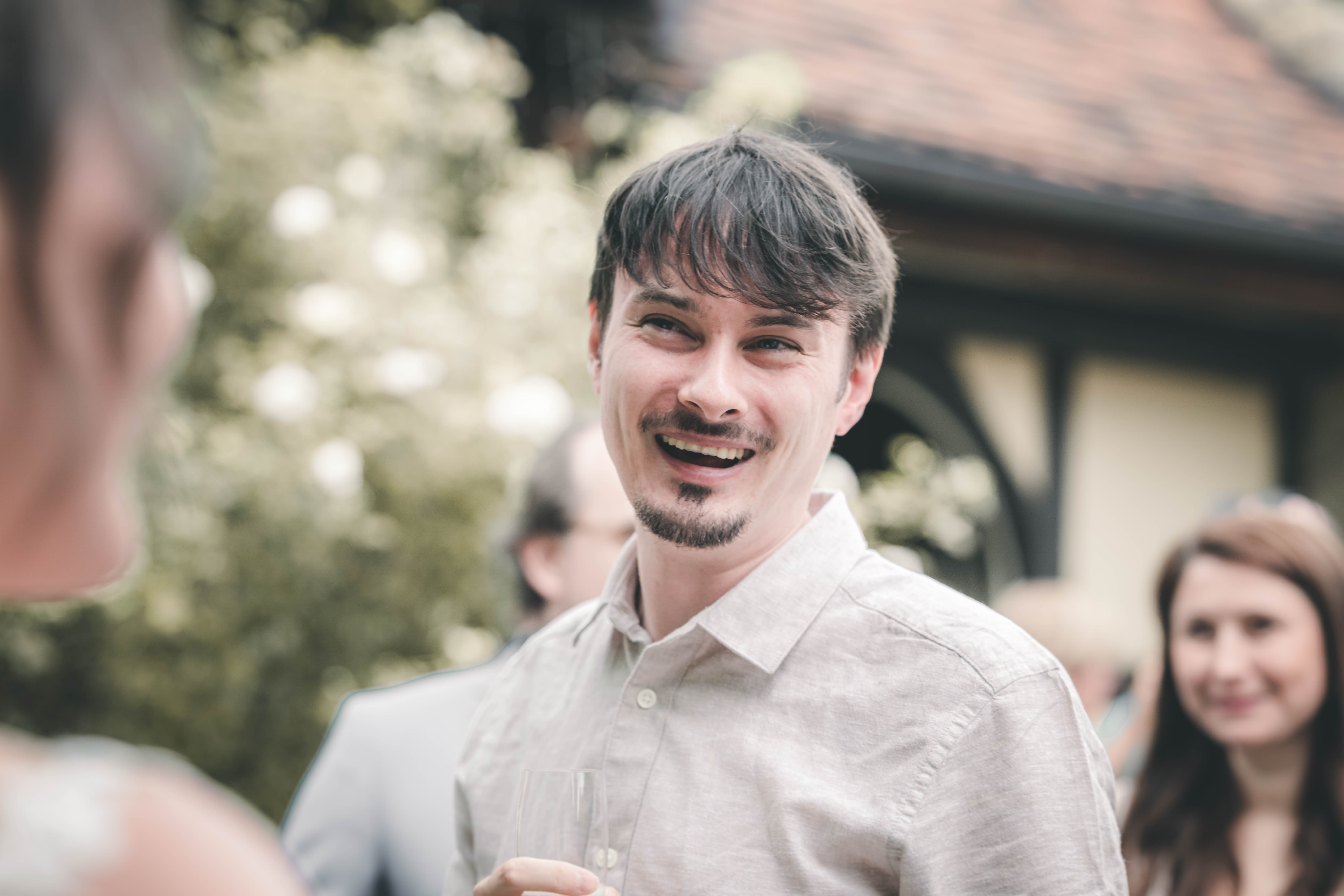 Ein junger Mann im Leinenhemd mit Spitzbart lacht mit einem Sektglas in der Hand bei der Hochzeitsfeier im Magdalenengut Erfurt.