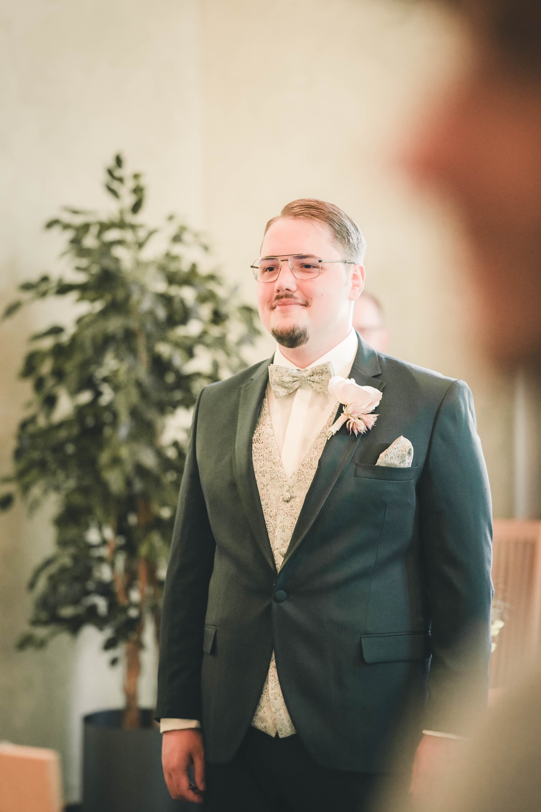 Der Bräutigam im eleganten dunkelgrünen Anzug mit floraler Weste, Fliege und Brille wartet im Standesamt Merseburg gespannt auf das Eintreffen seiner Braut.