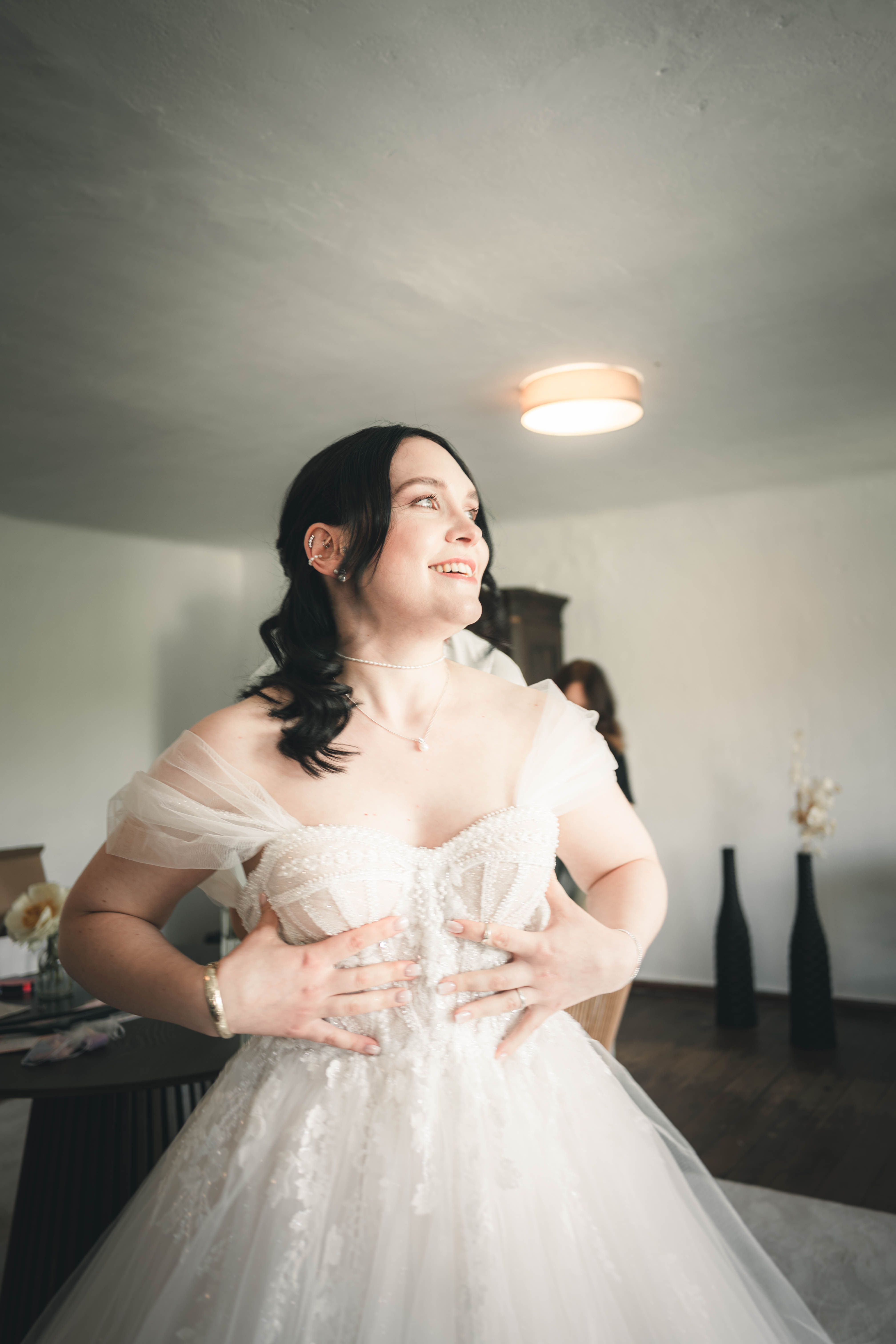 Die Braut blickt mit einem emotionalen, glücklichen Lächeln an sich herab, während sie die funkelnde Corsage ihres Hochzeitskleides richtet.