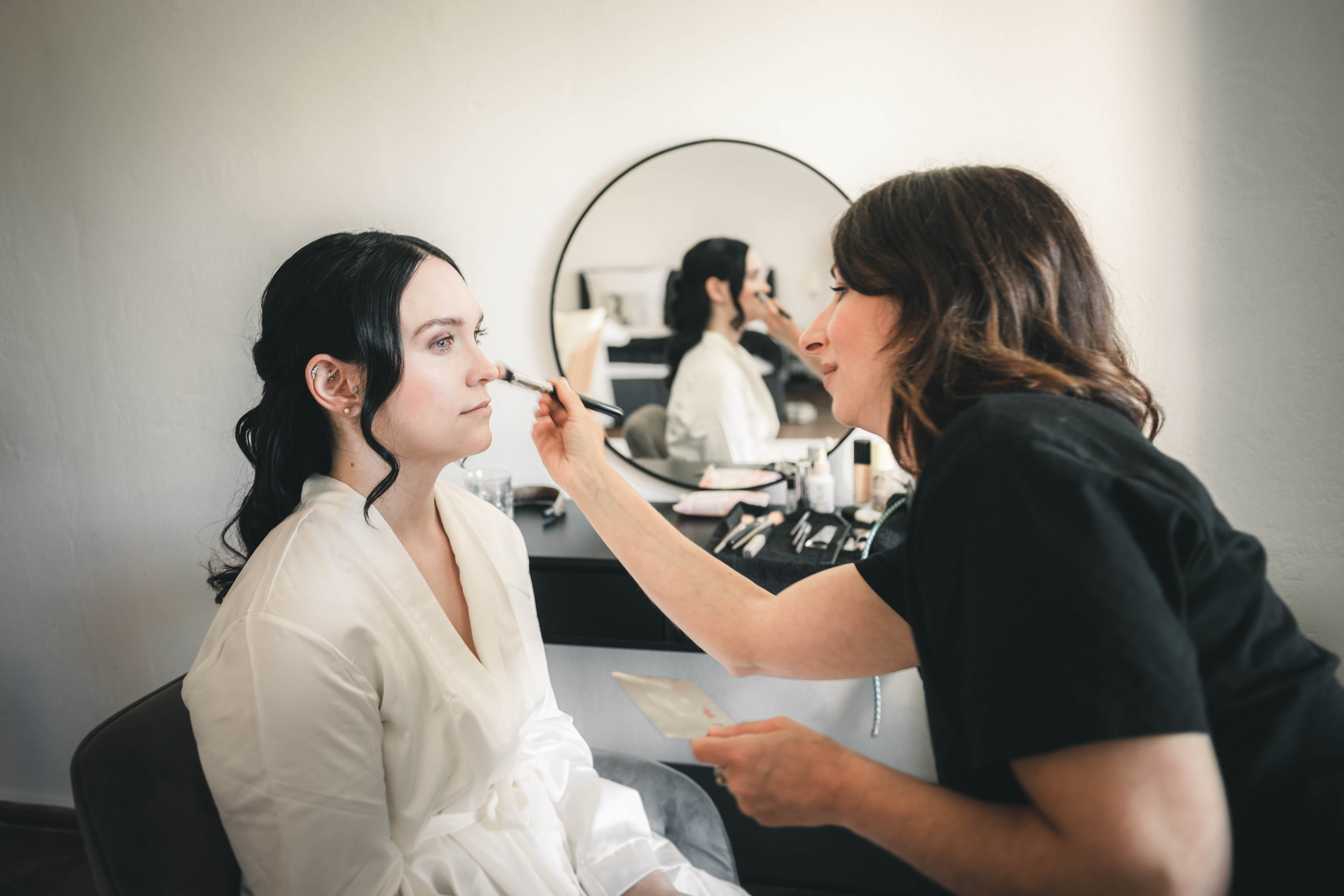 Die Make-up-Artistin trägt der Braut im weißen Morgenmantel mit einem Pinsel präzise das Make-up auf, im Hintergrund spiegelt sich die Szene im Rundspiegel.