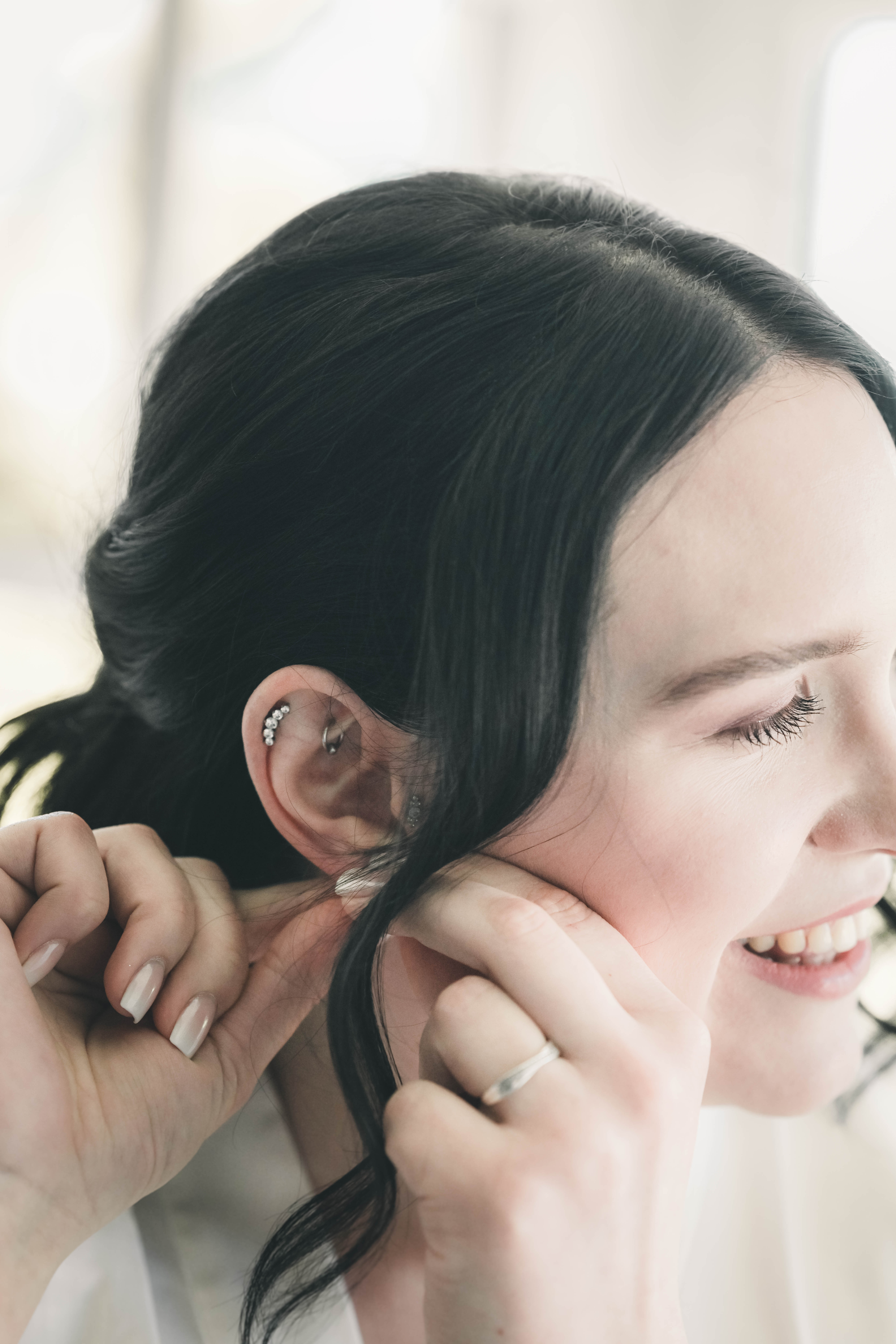 Detailnahe Aufnahme der lächelnden Braut im Profil, die ihre Haare richtet und ihren funkelnden Ohrschmuck sowie feine Piercings zeigt.