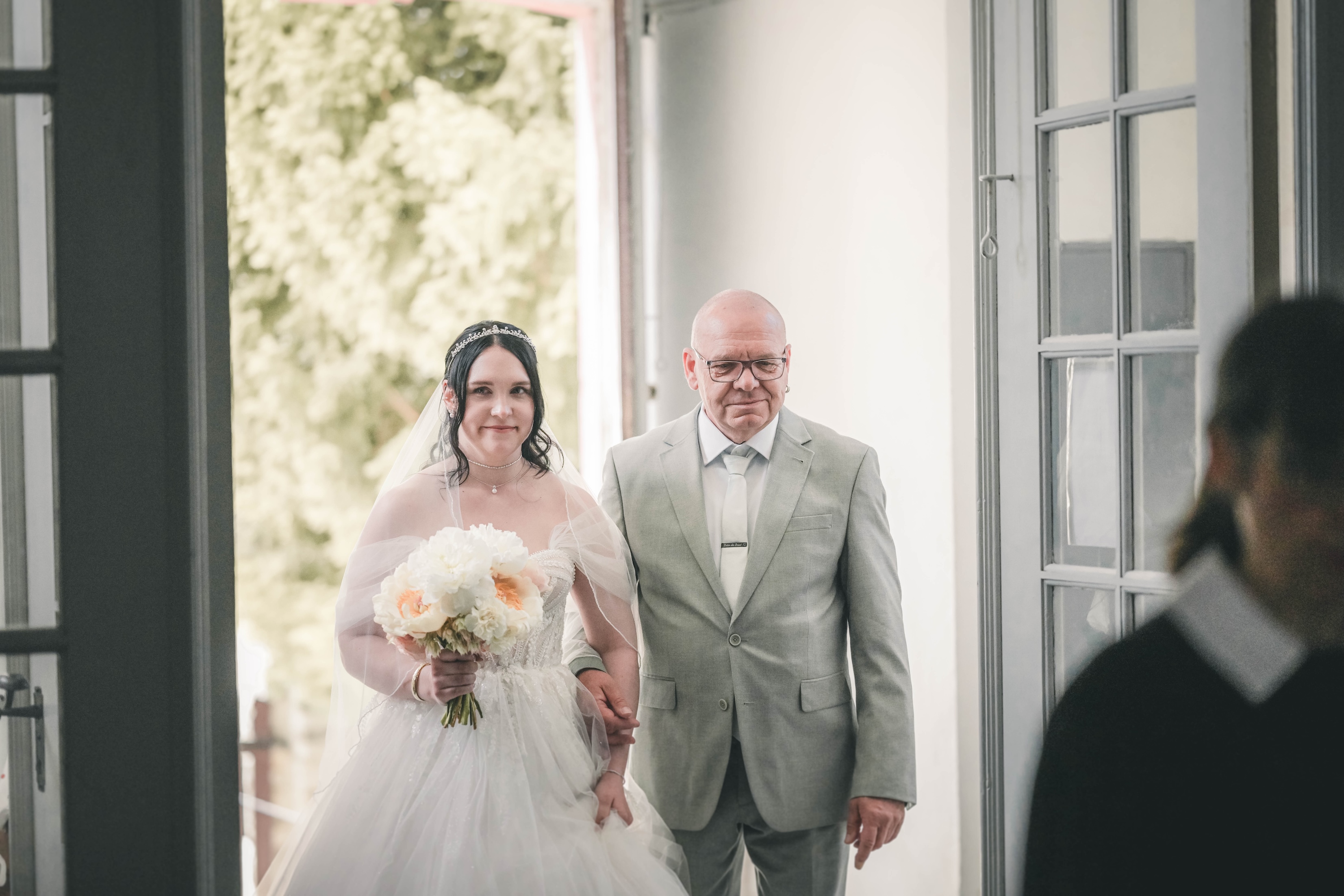 Der Brautvater führt seine sichtlich bewegte Tochter im Prinzessinnenkleid und mit dem Pfingstrosenstrauß im Arm durch die geöffnete Tür in die Kirche Frankleben.