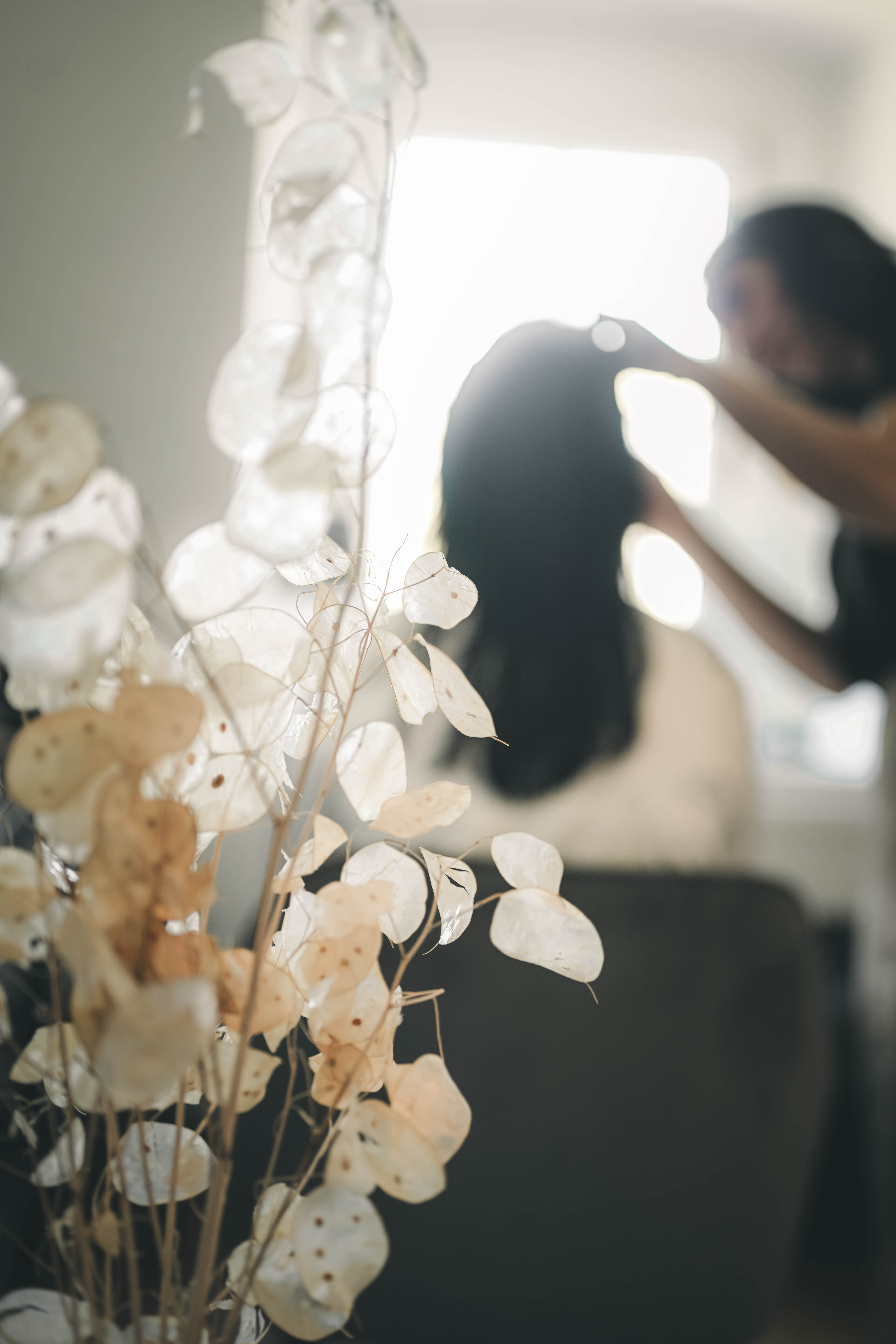 Eine kreative Aufnahme durch filigrane, helle Trockenblumen im Vordergrund auf die verschwommene Silhouette der Braut beim Haarstyling.