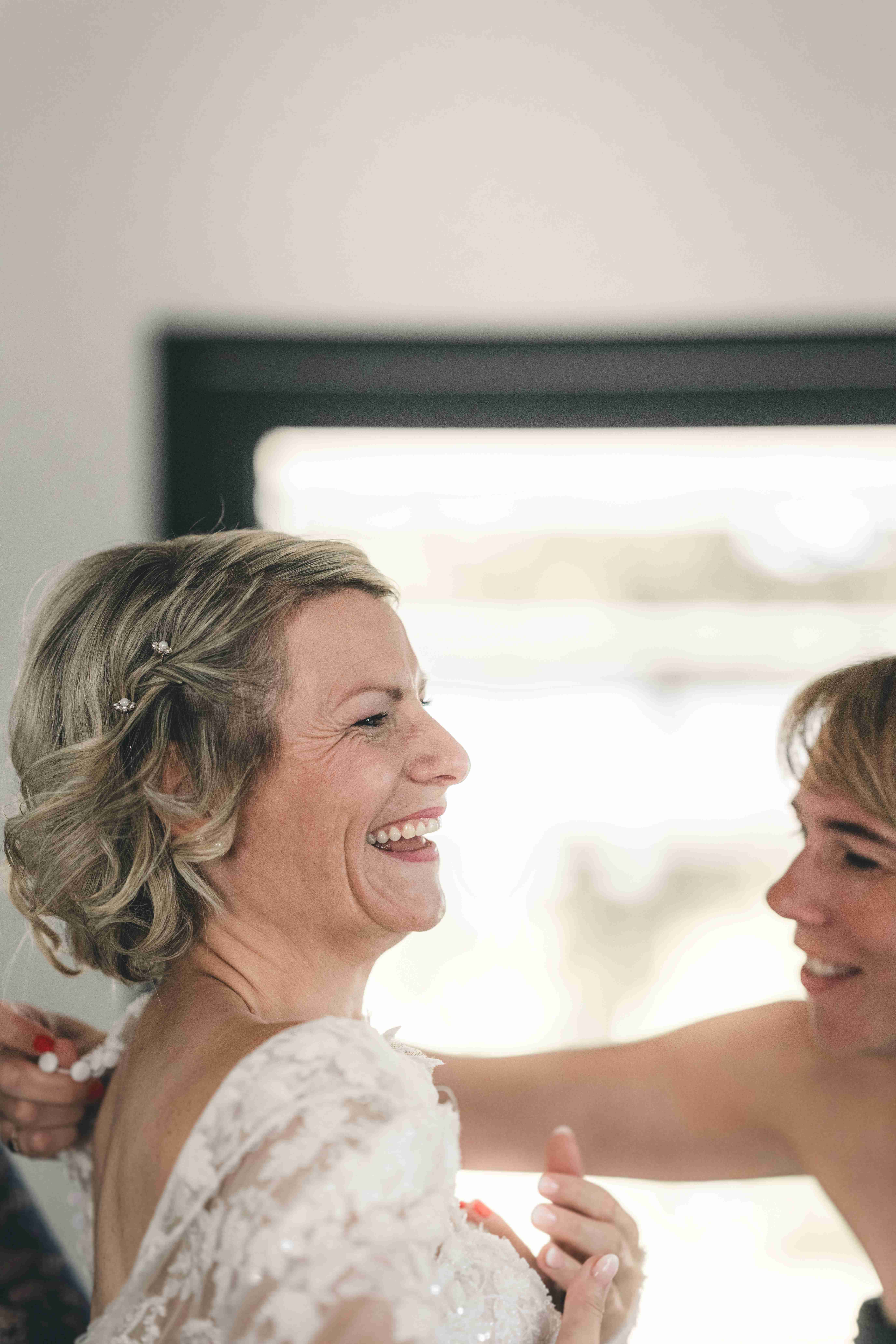 Ein emotionales Portrait der Braut, die sichtlich gerührt und glücklich auflacht, während ihr das Hochzeitskleid am Rücken geschlossen wird.