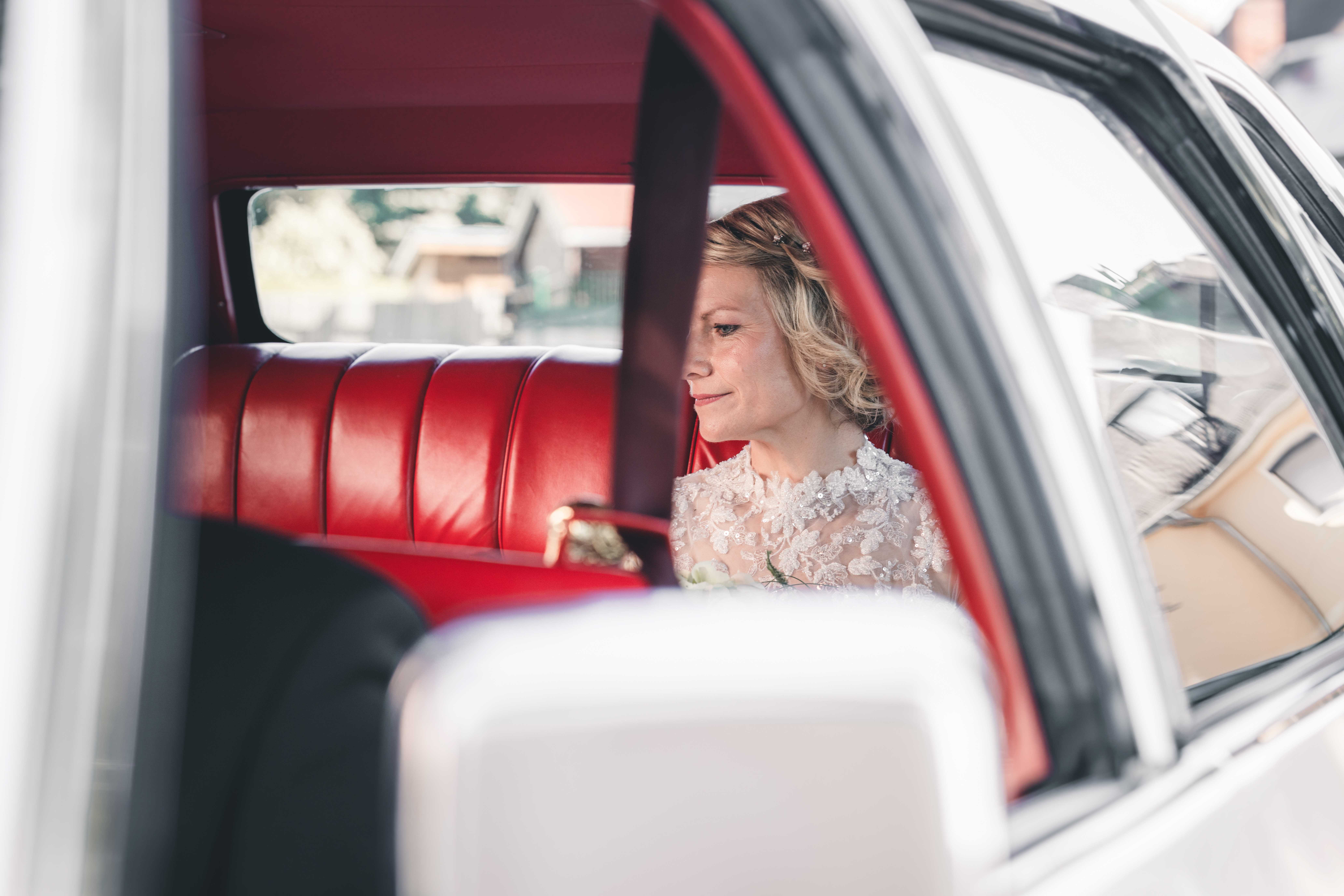 Ein Blick durch das Fenster des Oldtimers auf die Braut, die mit einem sanften Lächeln auf den roten Ledersitzen im Auto verweilt.