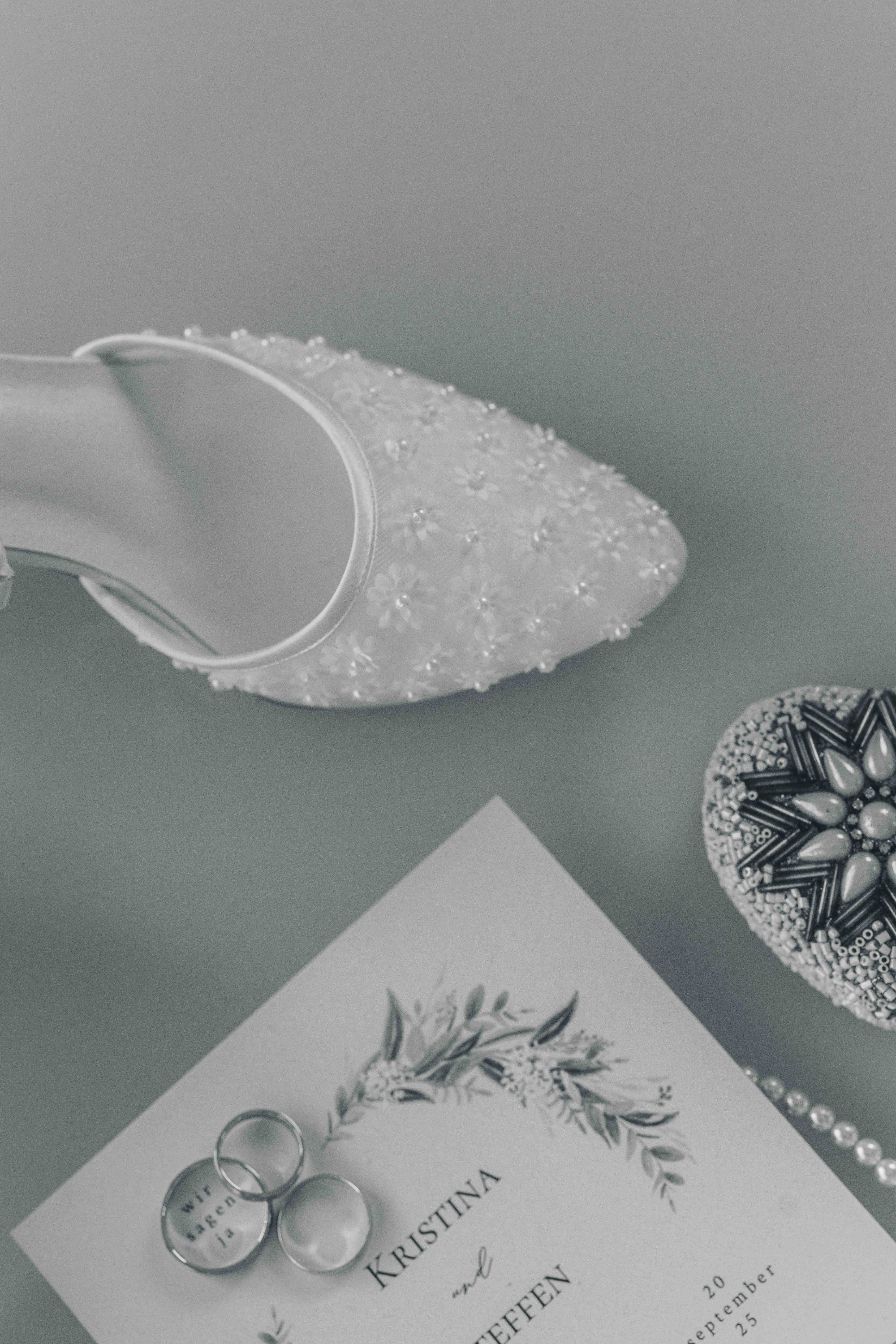 Eine kunstvolle Schwarz-Weiß-Nahaufnahme der gravierten Eheringe auf der Hochzeitseinladung neben einem eleganten Brautschuh mit Perlenbesatz.