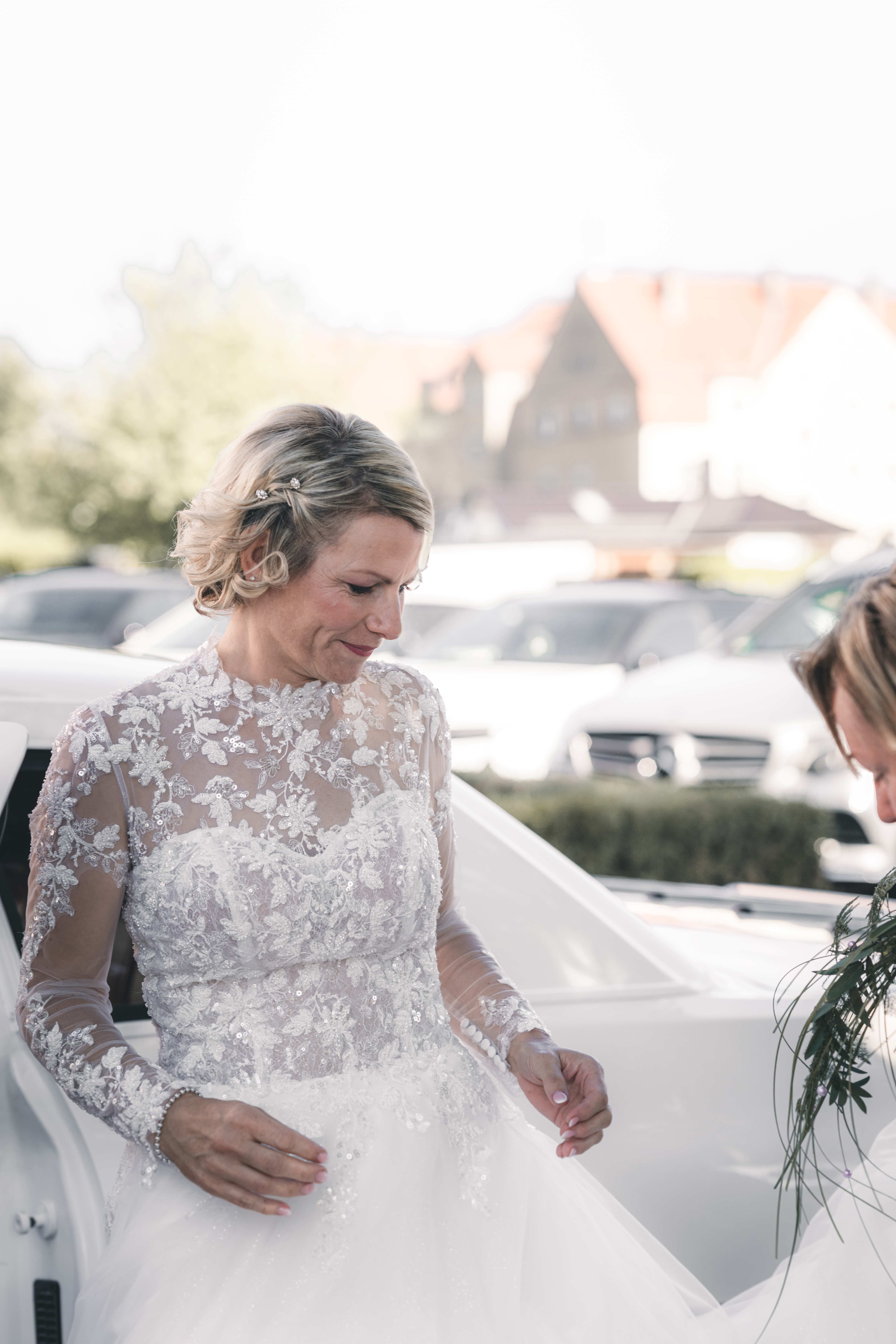 Die Braut im hochgeschlossenen Spitzenkleid steigt elegant aus dem weißen Hochzeitsauto vor der Traulocation im Erzgebirge.