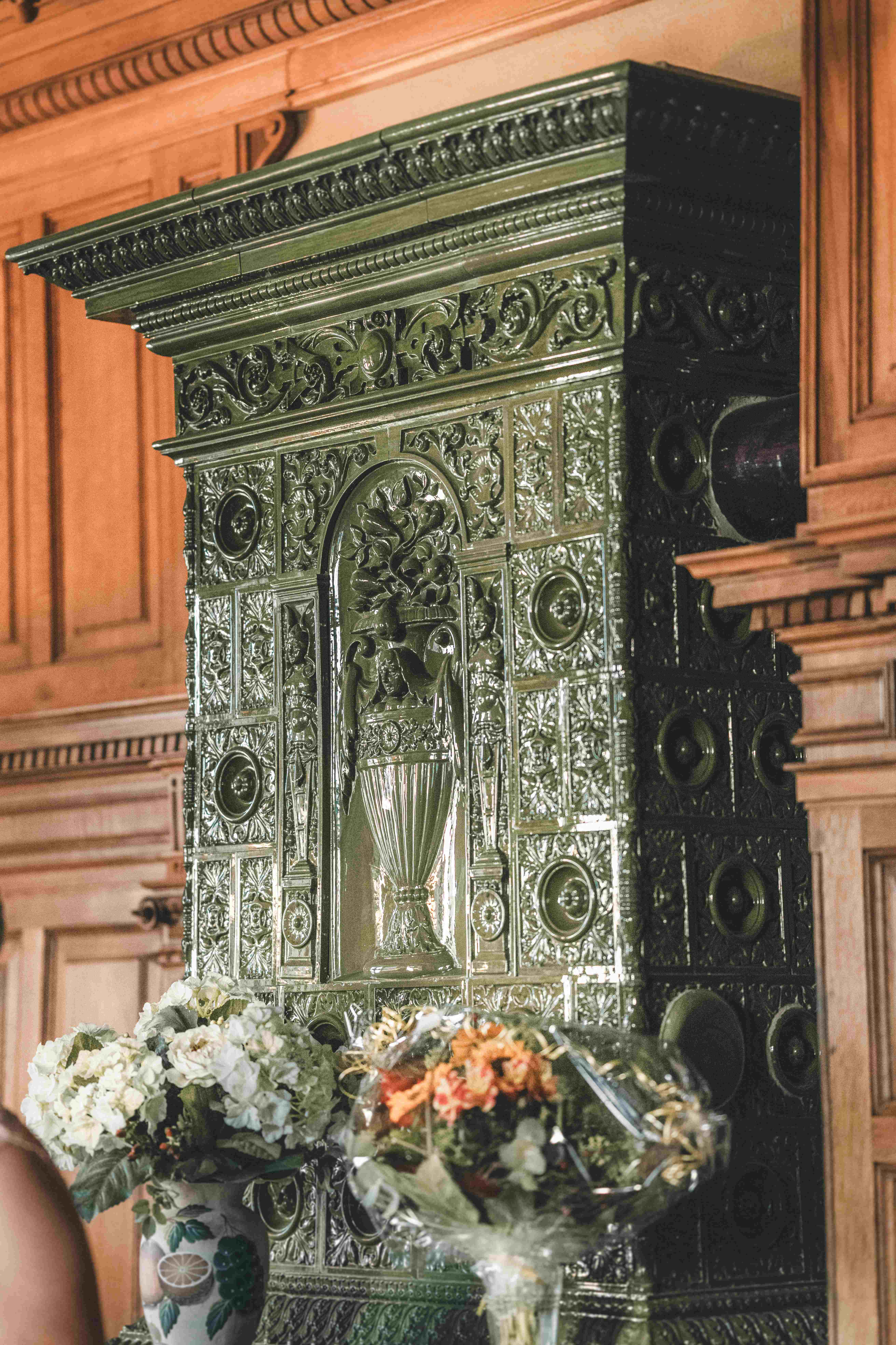 Ein detailreich verzierter, historischer grüner Kachelofen im Trausaal des Herrenhauses Fraureuth, geschmückt mit Hochzeitsblumen.